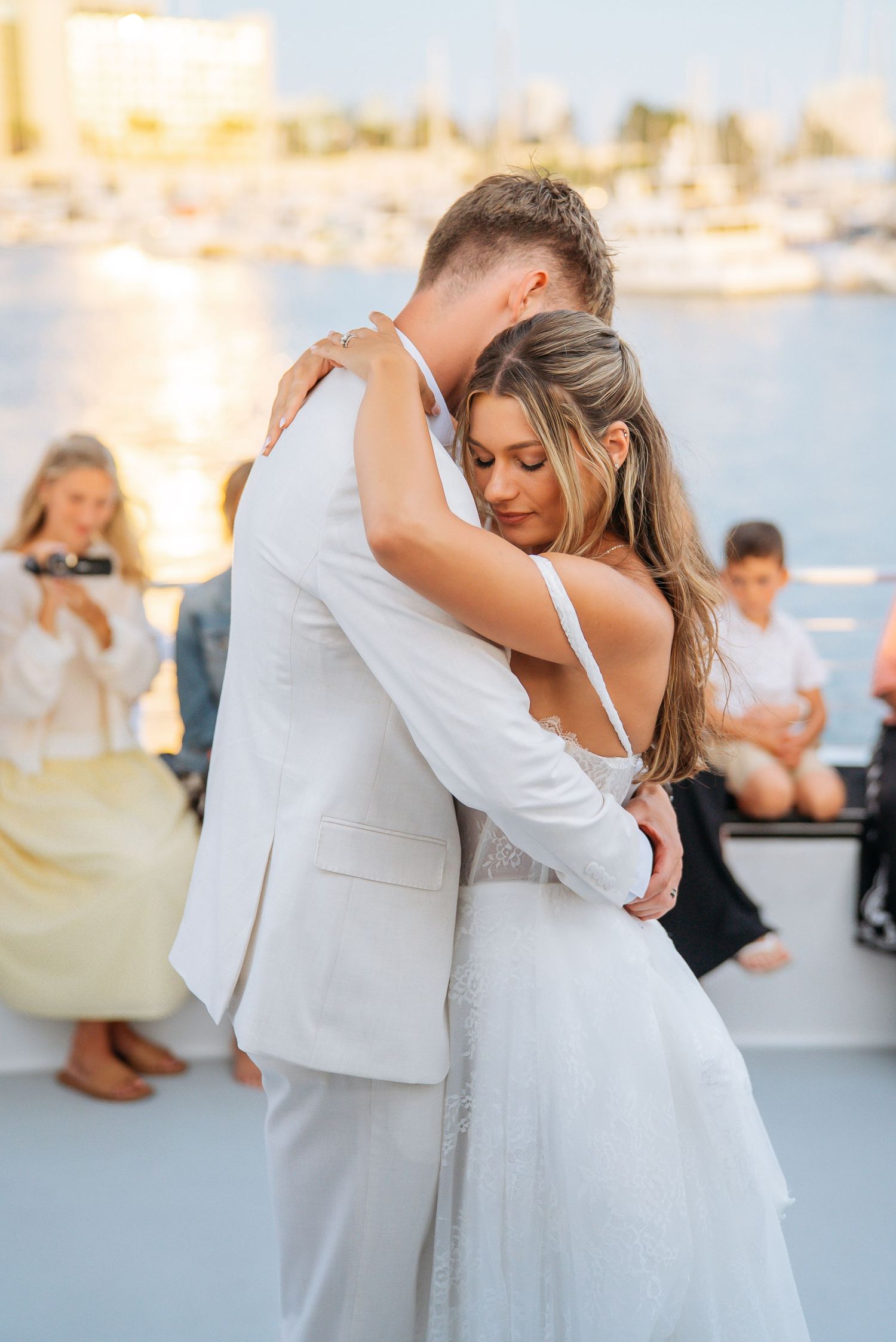 Bride and groom share their first dance on a yacht at sunset, guests watching in the background.