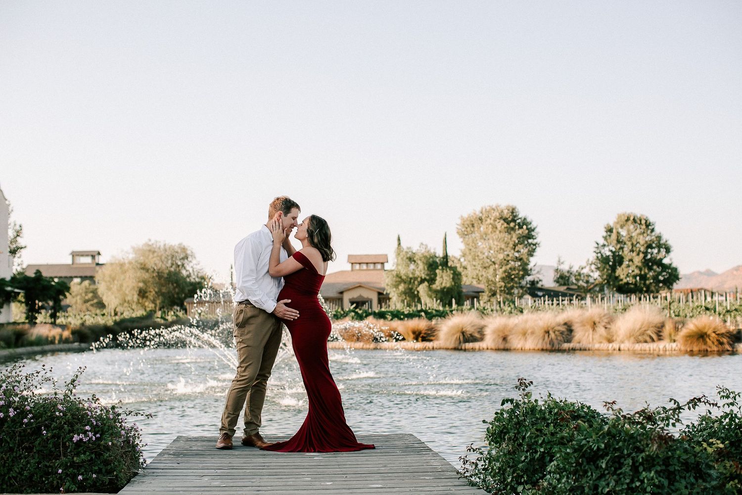Couple embracing on a dock by a fountain, woman in a red gown, man in white shirt, at a scenic outdoor venue.