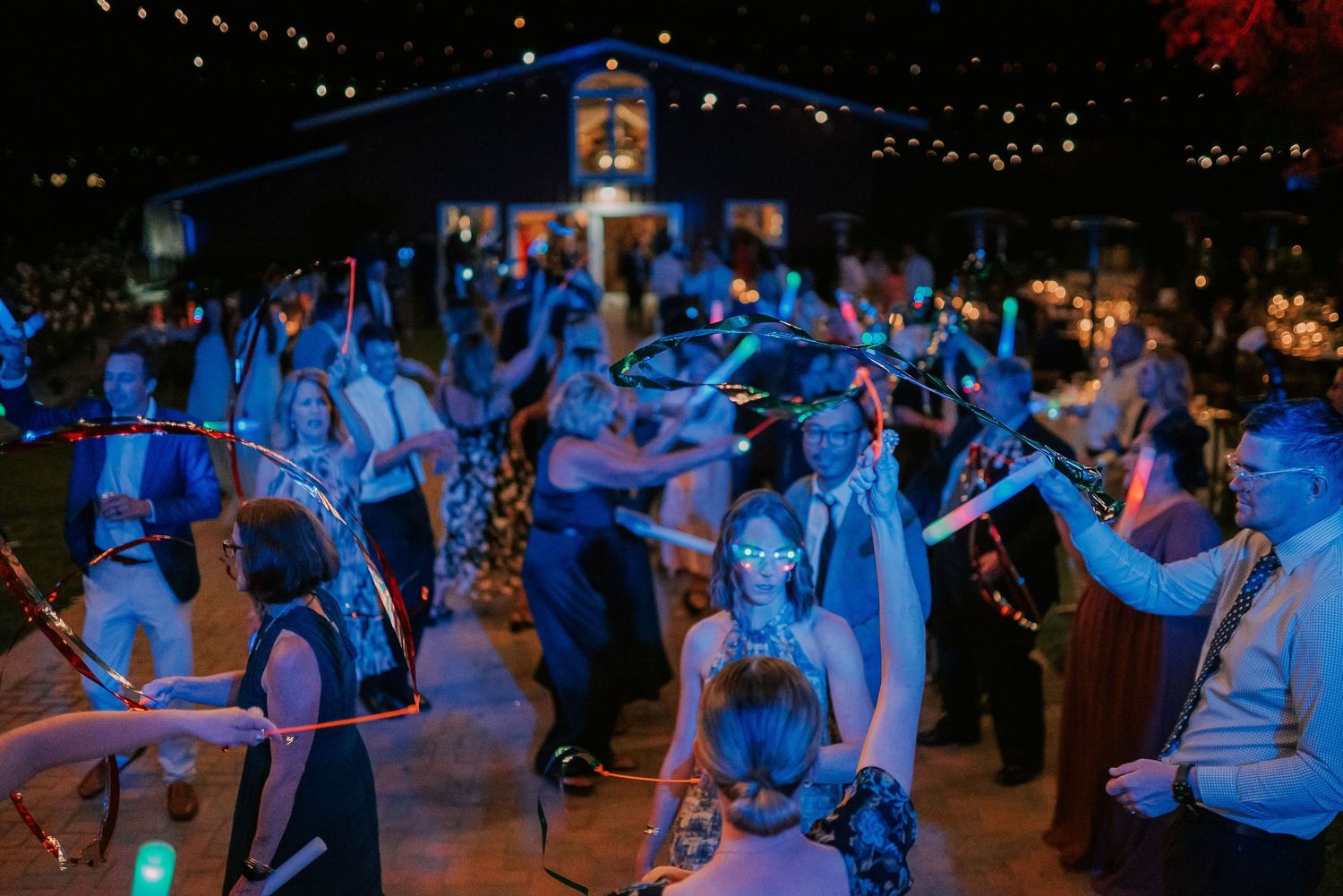 Guests dancing at night wedding reception with glowing neon hula hoops and string lights illuminating outdoor venue.