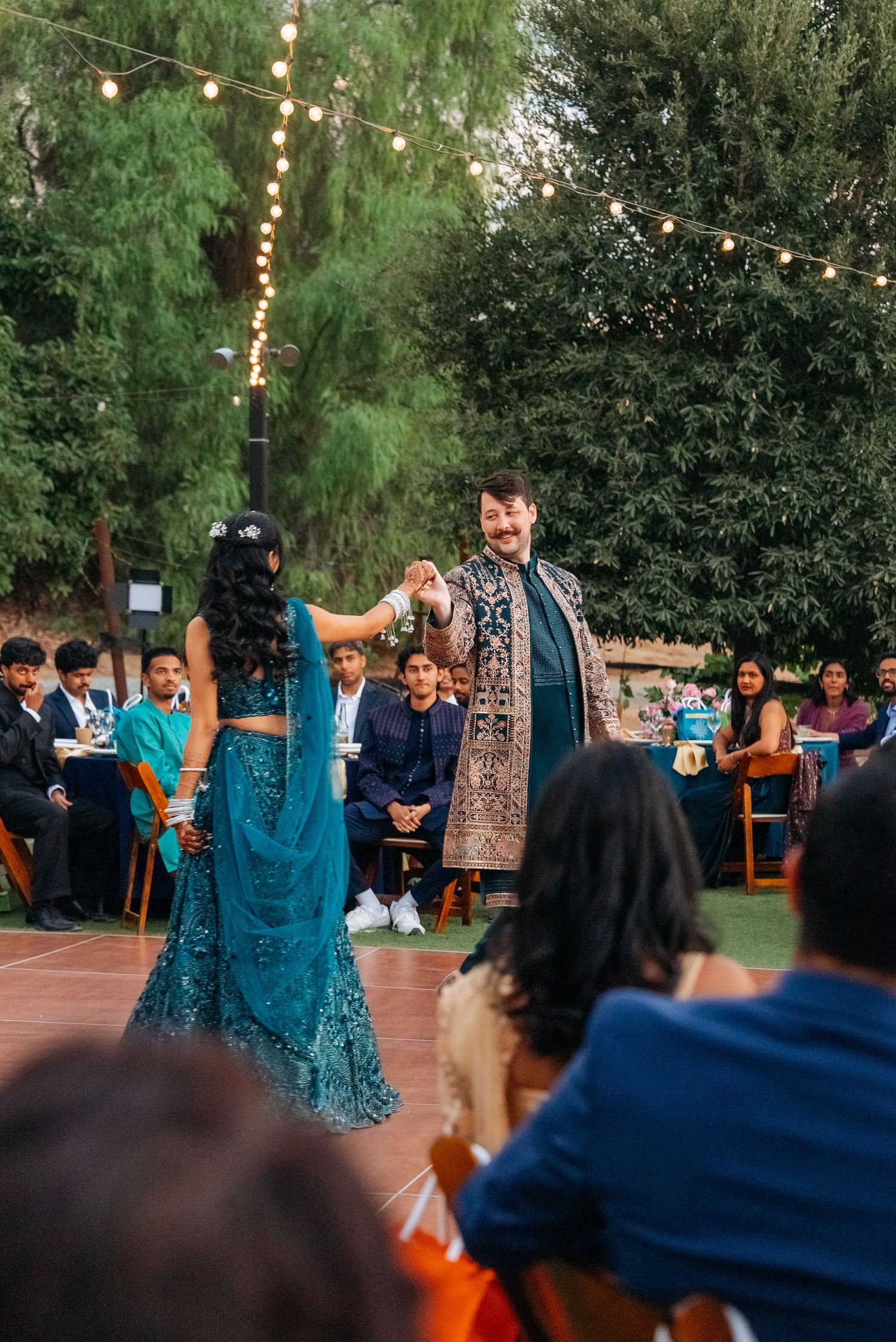 Couple in traditional Indian attire dancing at an outdoor wedding reception with string lights and seated guests.