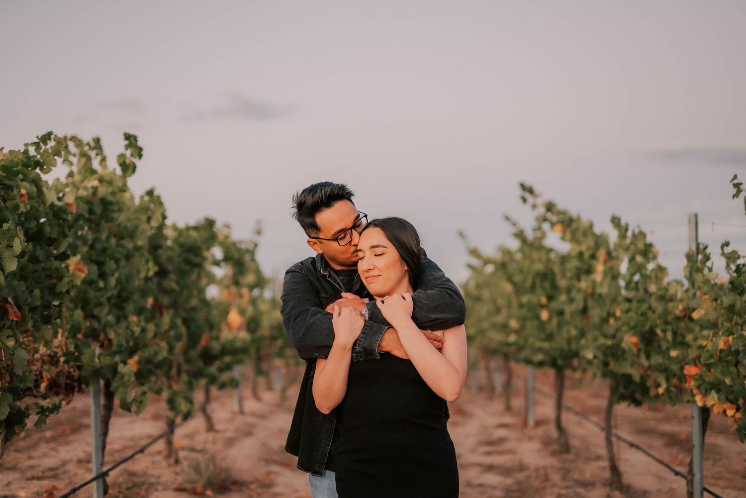 Couple embracing romantically in a vineyard at sunset, woman leaning back against man in black outfits.