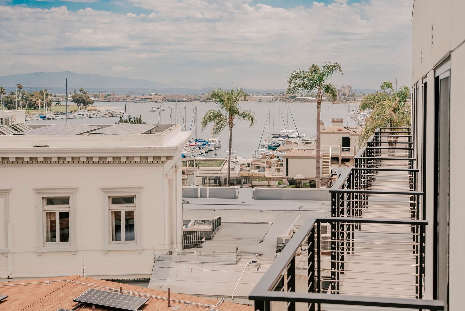 Rooftop balcony view overlooking a sunny marina with sailboats, palm trees, and white buildings under a partly cloudy sky.