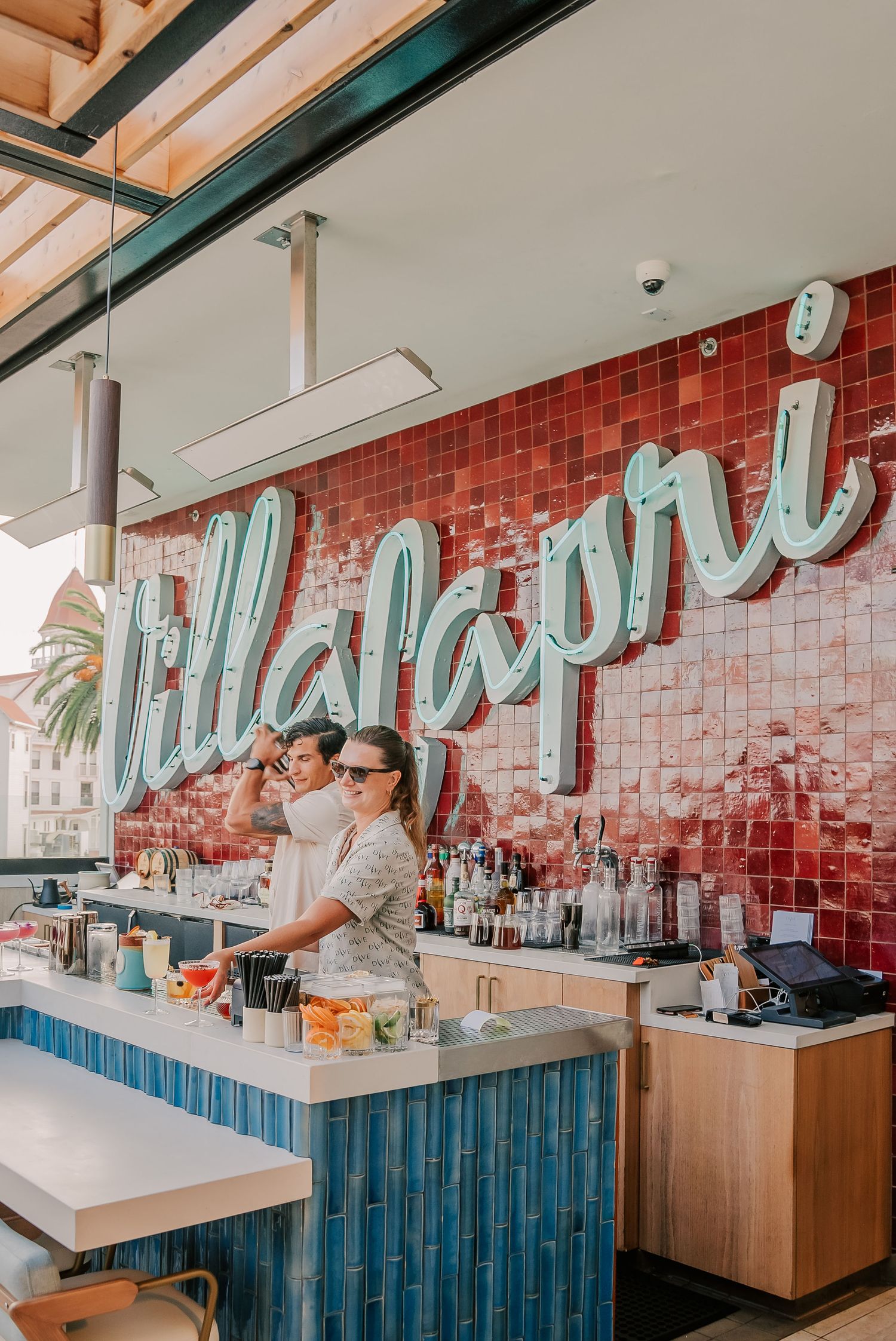 Two bartenders work behind the bar at Villa Capri restaurant, featuring a red tile wall with cursive neon signage.