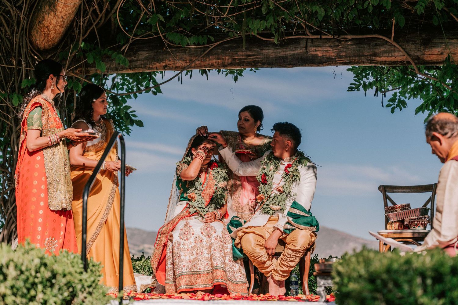 Traditional Indian wedding ceremony with bride and groom seated under decorated mandap outdoors.