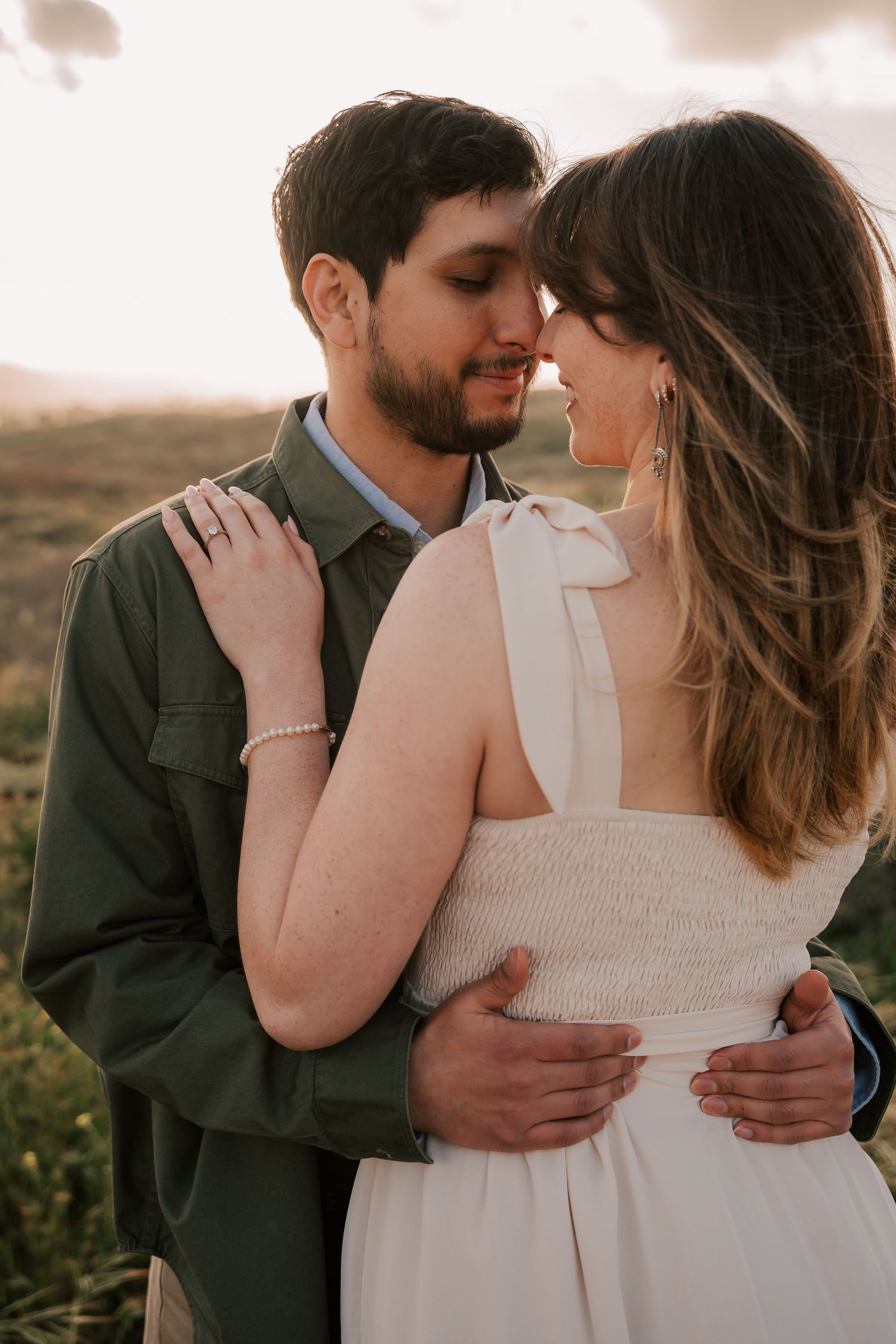 Couple embracing outdoors at sunset, a bearded man in an olive green jacket holding a woman in a white sundress with long hair.