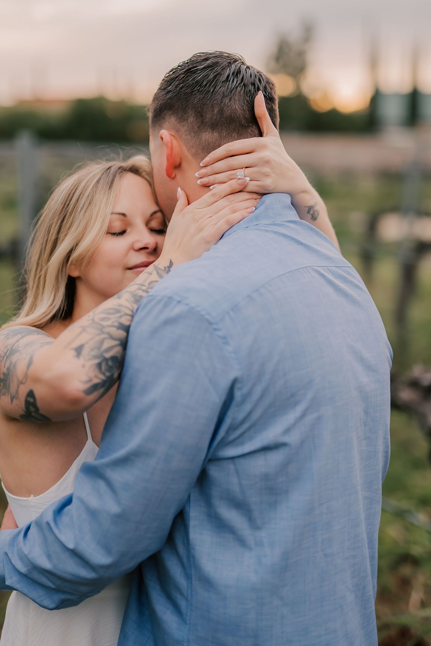 A couple embraces tenderly at sunset in a vineyard, the woman with tattoos smiling peacefully against her partner's shoulder.