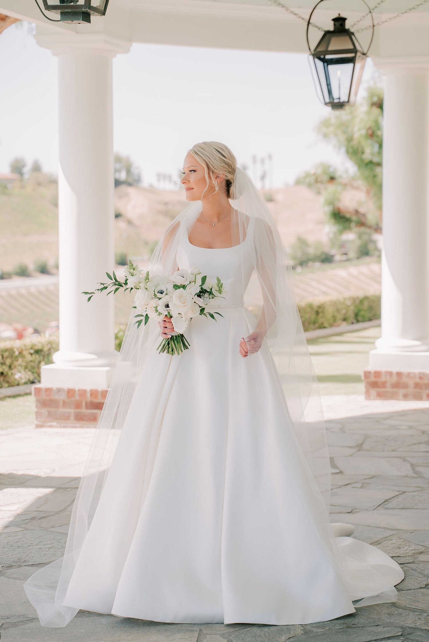 Bride in white ball gown with square neckline and veil holds white floral bouquet beside elegant columns.
