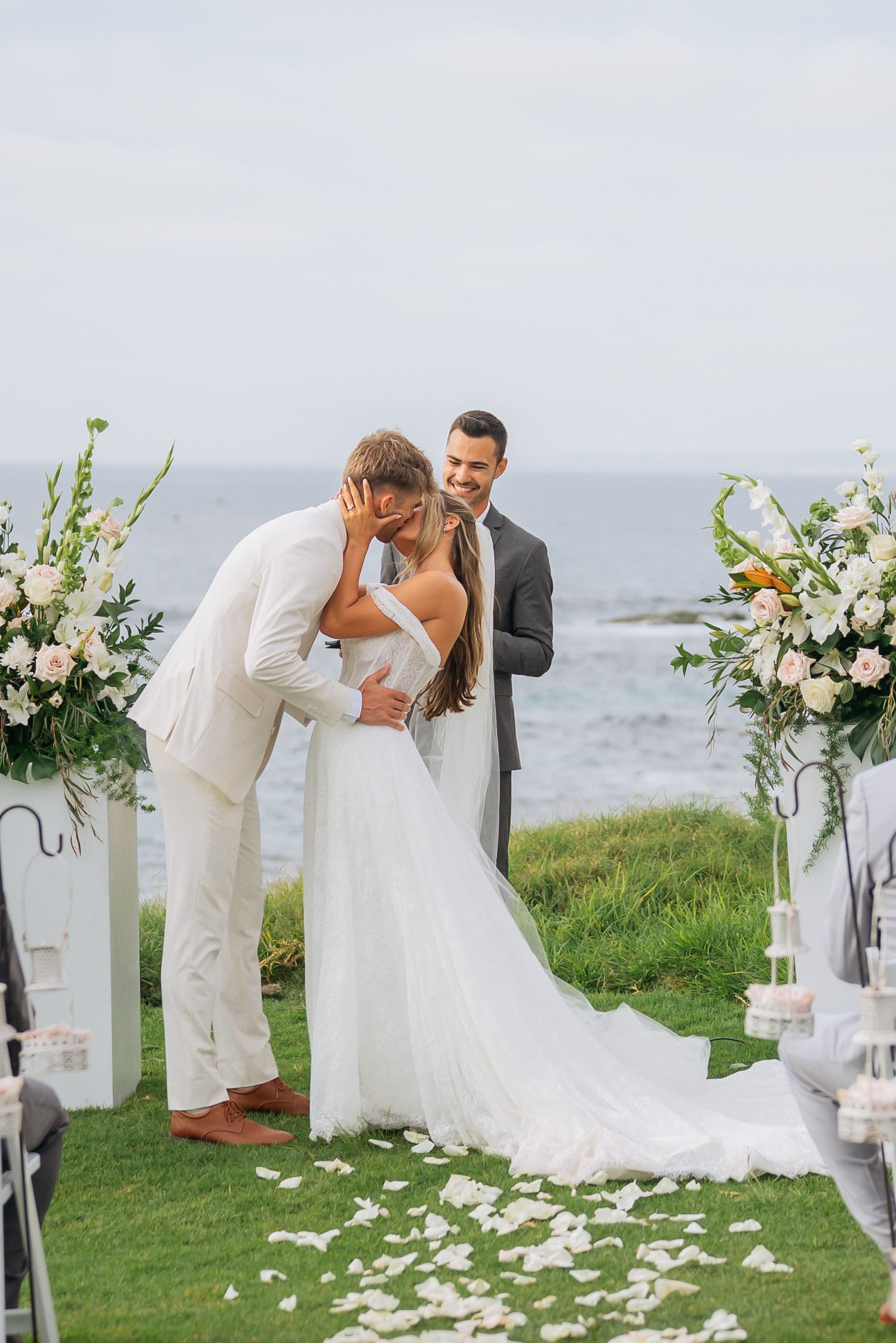 Bride and groom share first kiss at oceanside altar surrounded by white rose petals on grass.