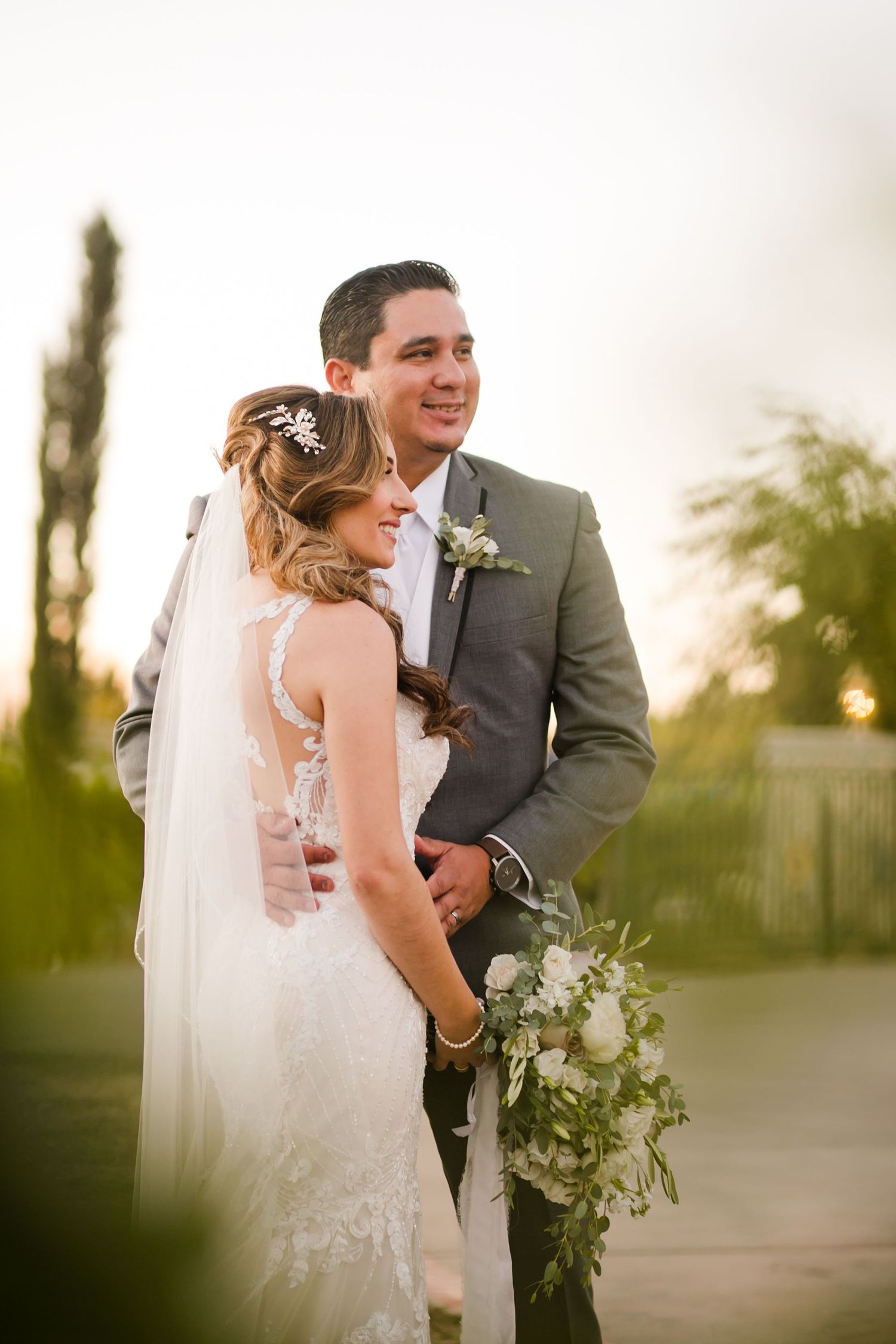 Bride in lace gown with veil and groom in gray suit embrace outdoors, holding white floral bouquet at golden hour.
