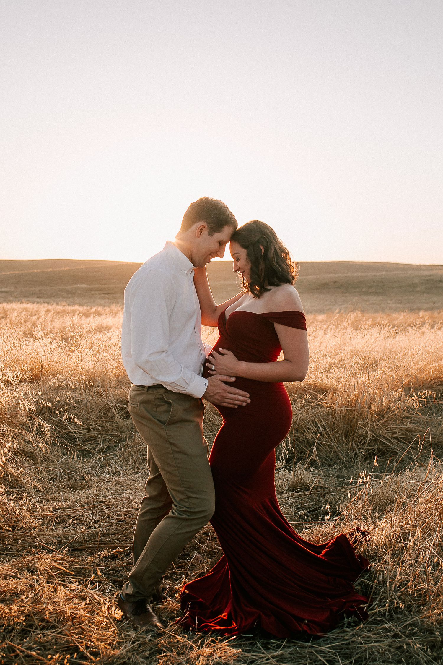 Couple embracing in golden field at sunset, woman in elegant red gown, romantic maternity photo session outdoors.