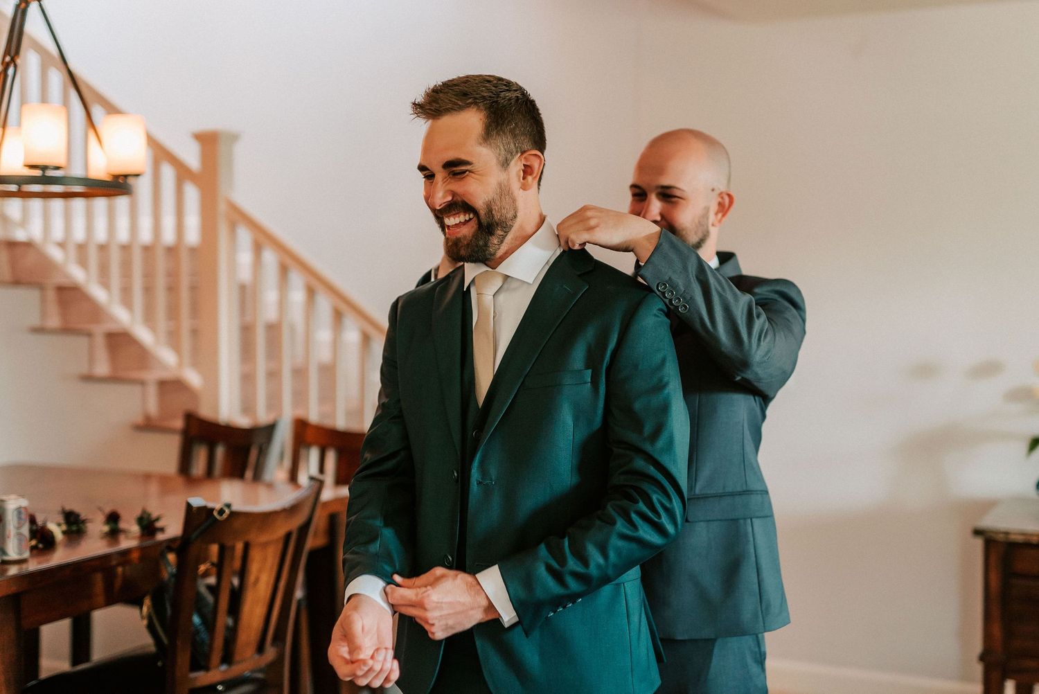 Groom smiling in green suit being helped by groomsman adjusting collar in elegant home staircase setting.