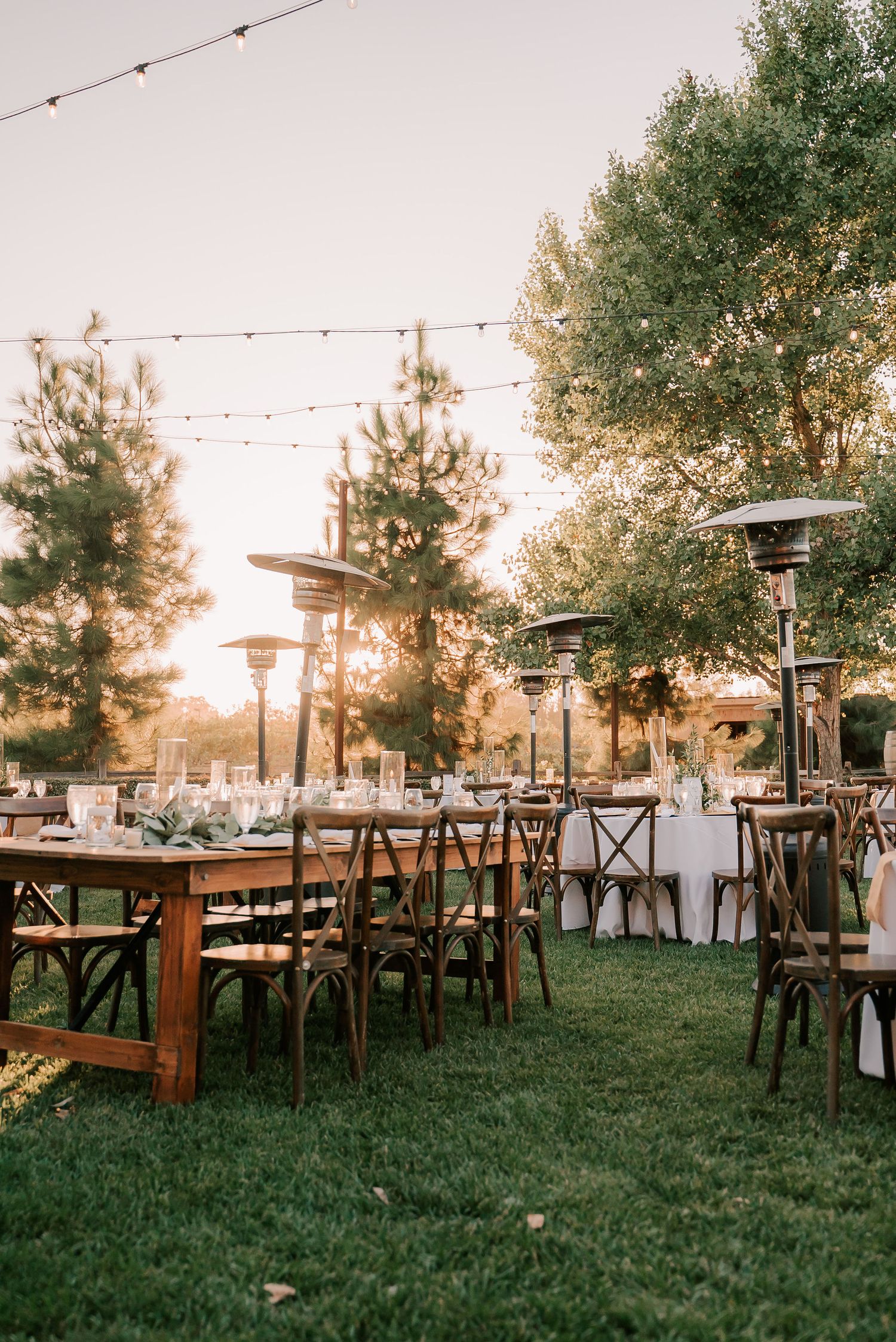 Outdoor wedding reception at sunset with rustic wood tables, string lights, and patio heaters on a lush green lawn.