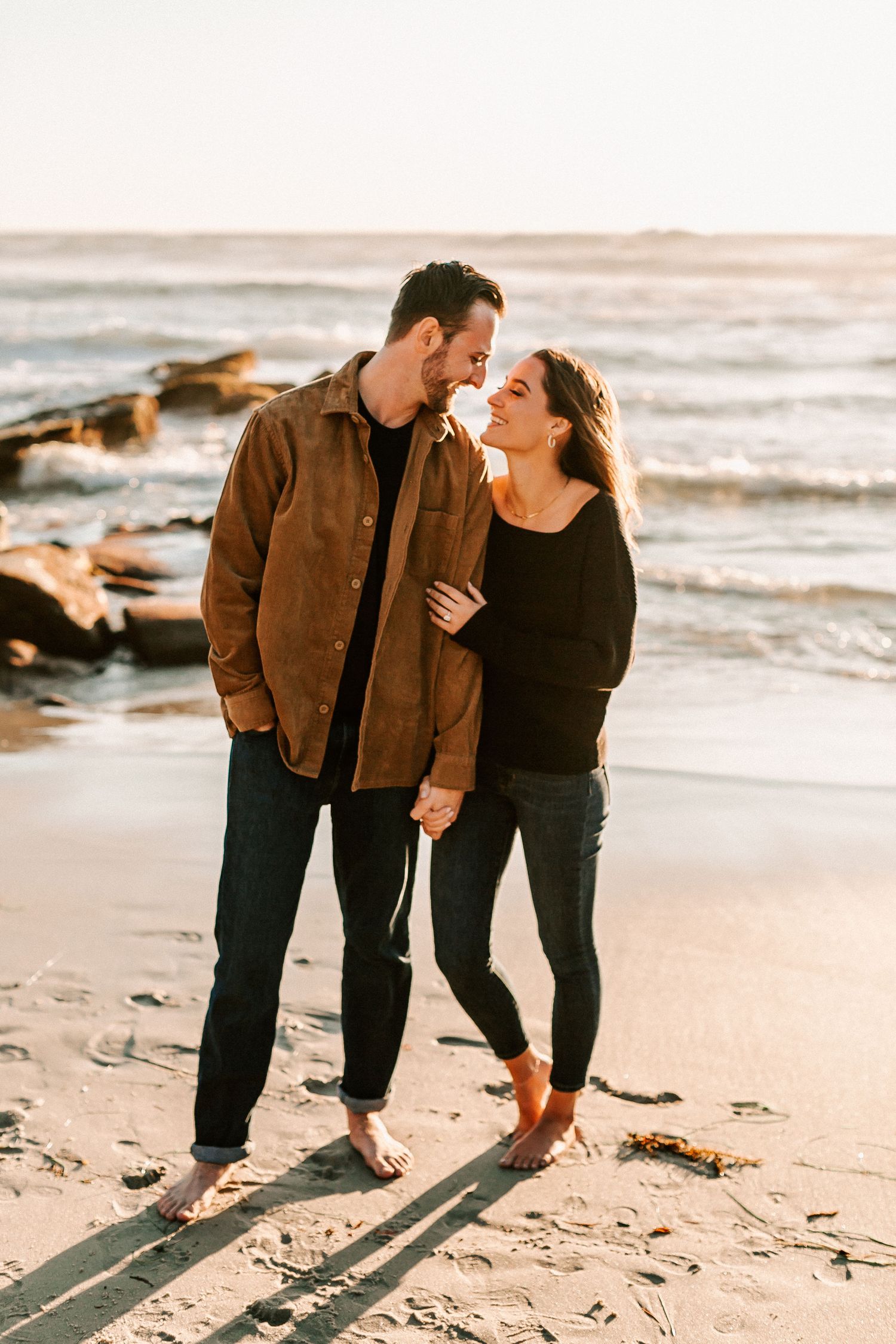 Barefoot couple gazing at each other on sandy beach at sunset, holding hands romantically.