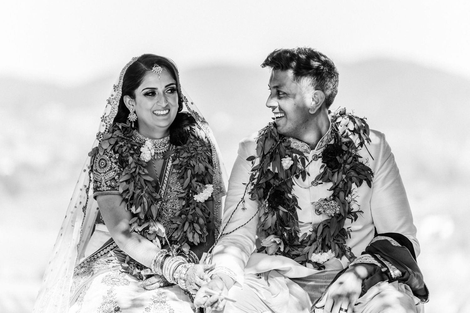 Black and white portrait of smiling Indian bride and groom with floral garlands.