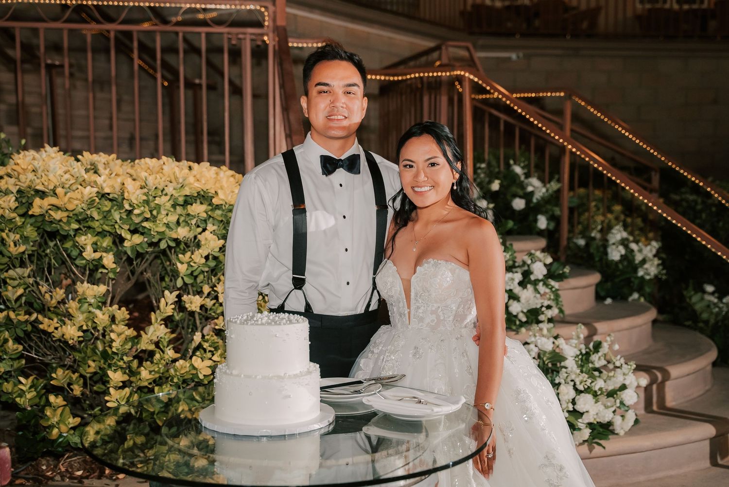 Bride and groom posing together beside a two-tier white wedding cake at an elegant outdoor reception venue.