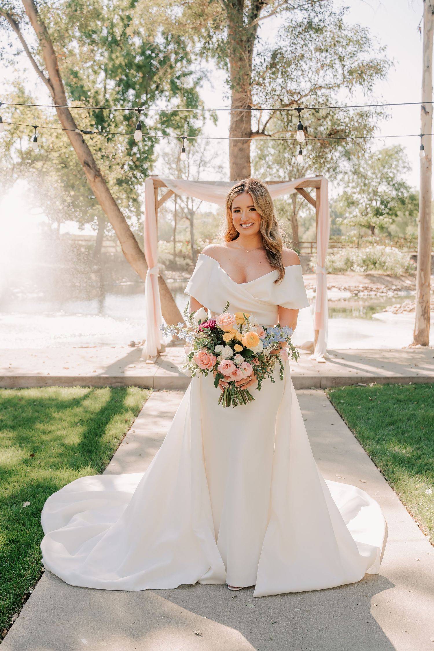 Bride in off-shoulder white gown holds colorful bouquet outdoors near wooden arch with greenery.