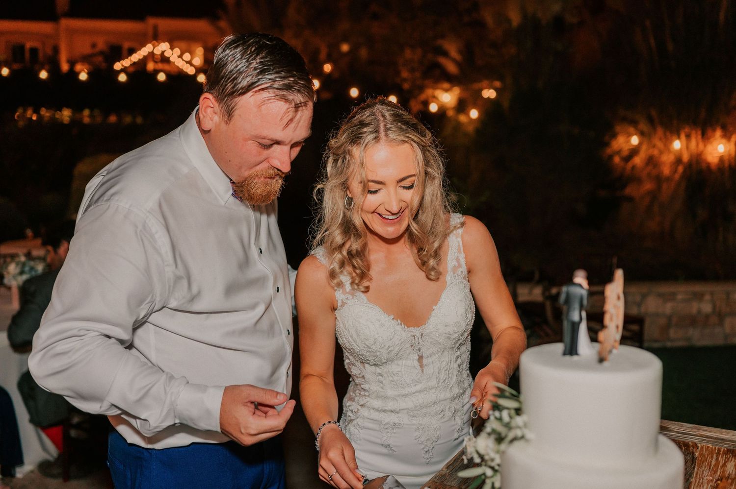 Bride and groom cutting white wedding cake together at outdoor evening reception with string lights.