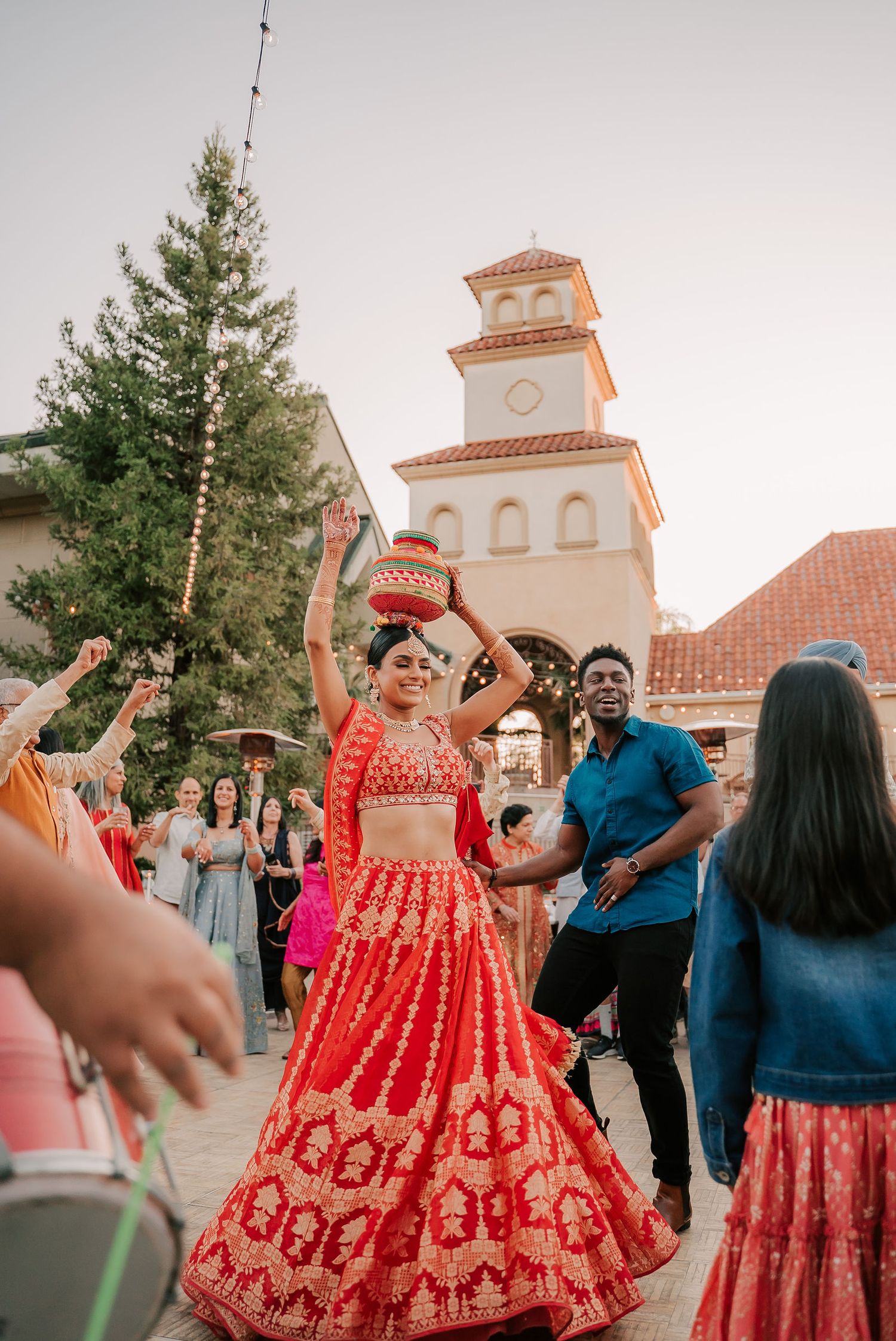 Indian bride in red lehenga balances pot on head while dancing at outdoor wedding celebration at dusk.