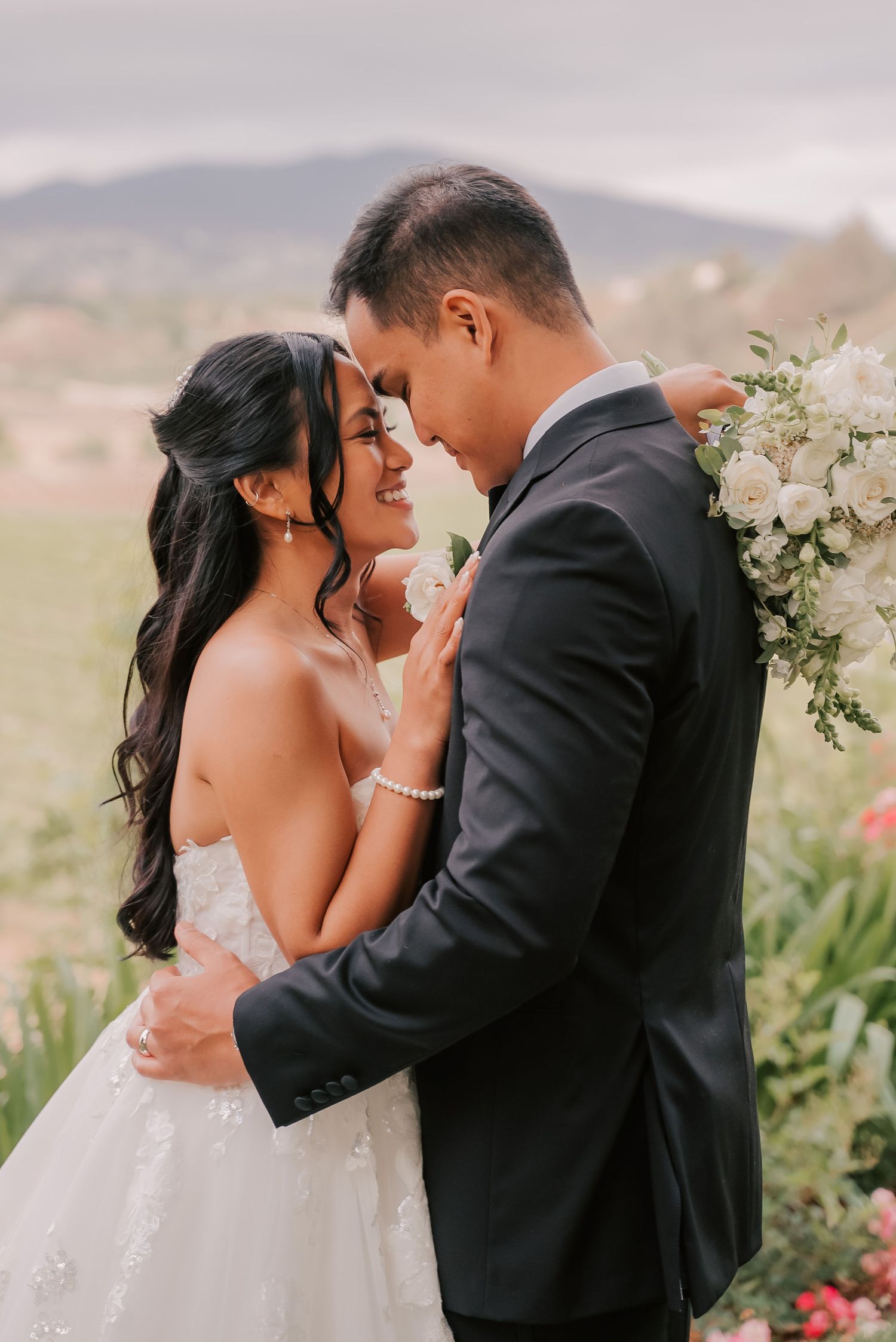 Bride and groom share a romantic embrace outdoors, smiling closely with white floral arrangement and vineyard backdrop.