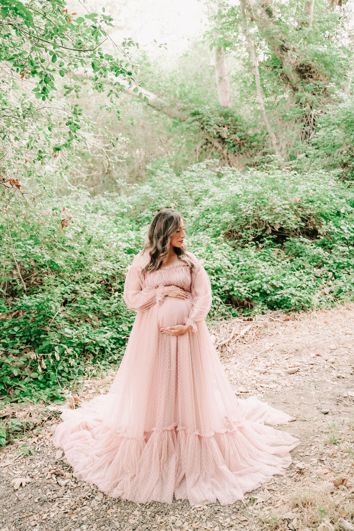 Pregnant woman in flowing pink tulle gown cradling baby bump outdoors in lush green forest setting.