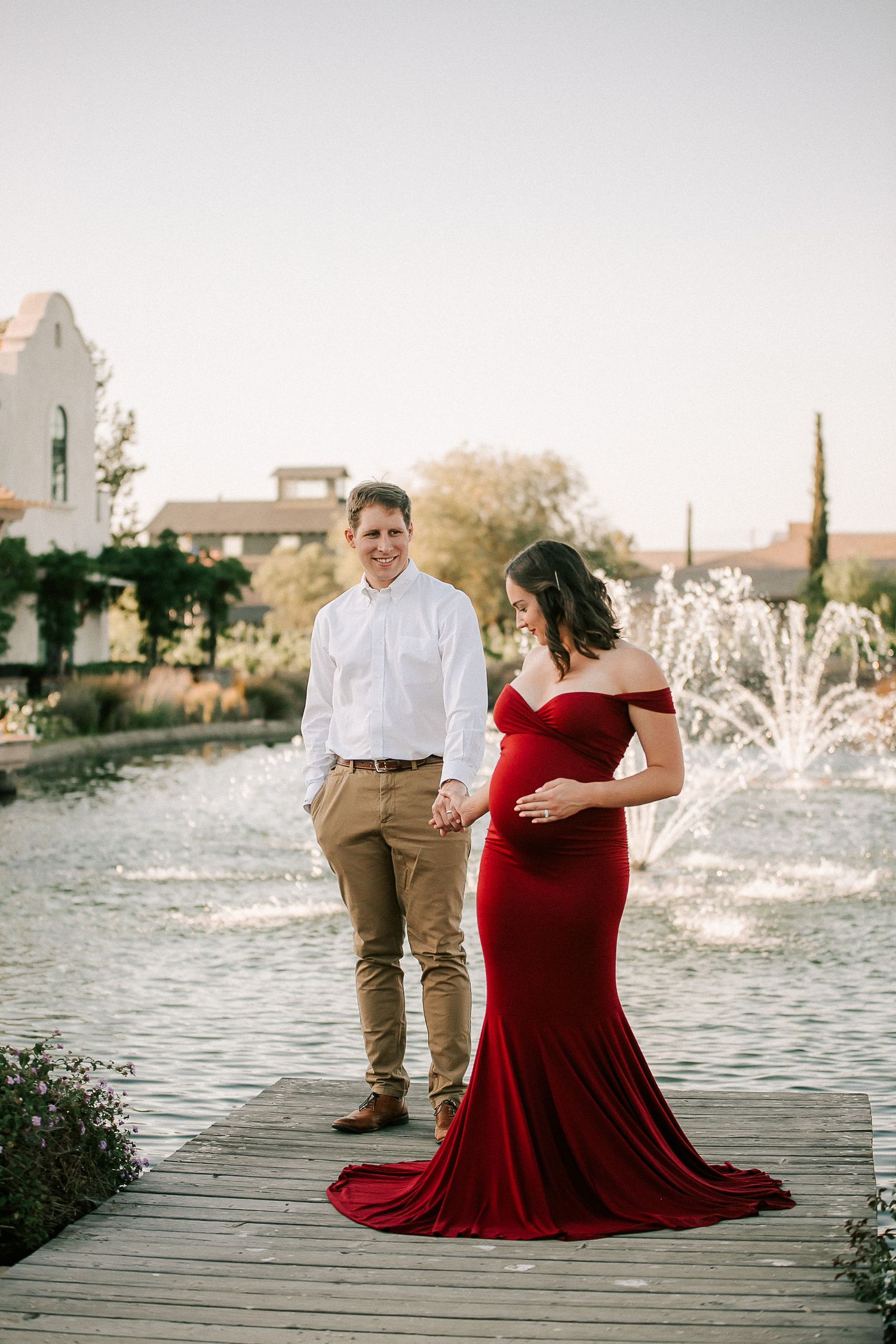 Pregnant woman in red gown holds belly near fountain, man in white shirt smiles beside her.
