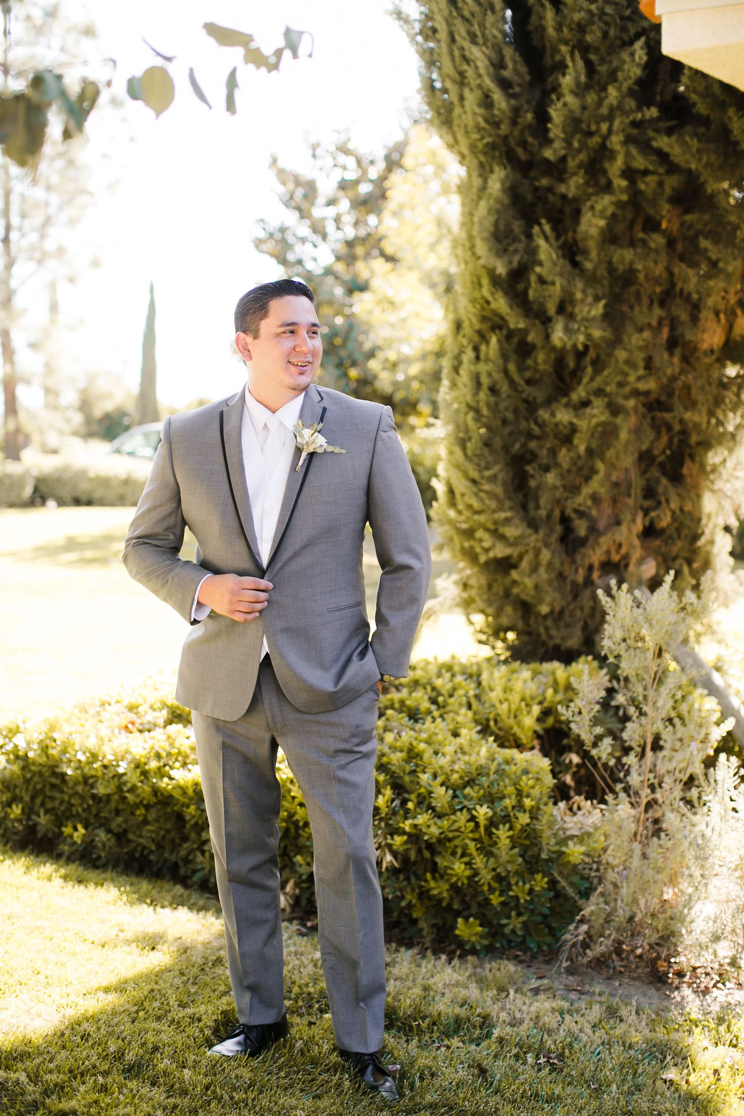 Smiling groom in gray suit with boutonniere standing outdoors on green lawn beside tall cypress trees.
