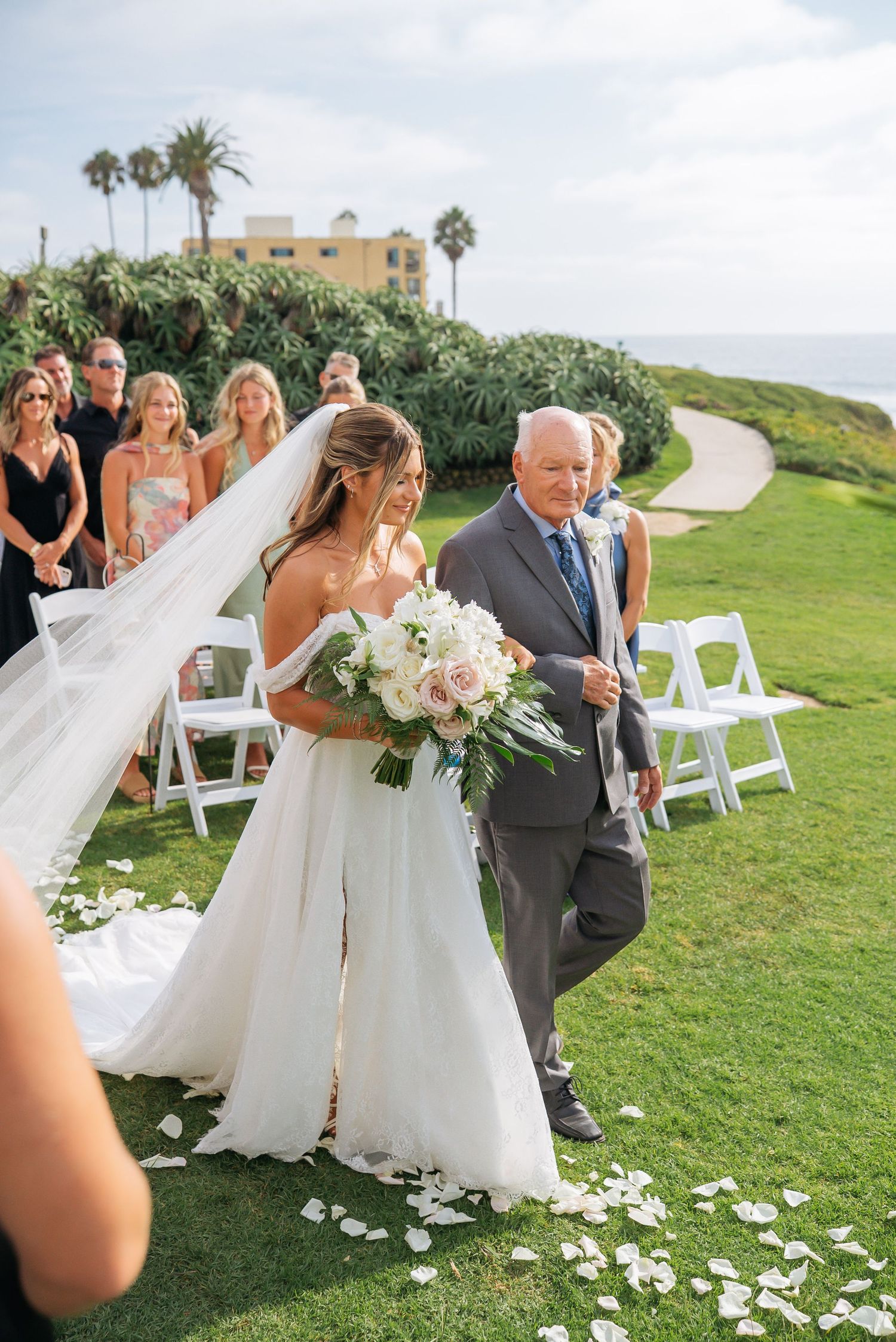 Bride walks down outdoor aisle with escort, white bouquet in hand, guests seated on sunny lawn.