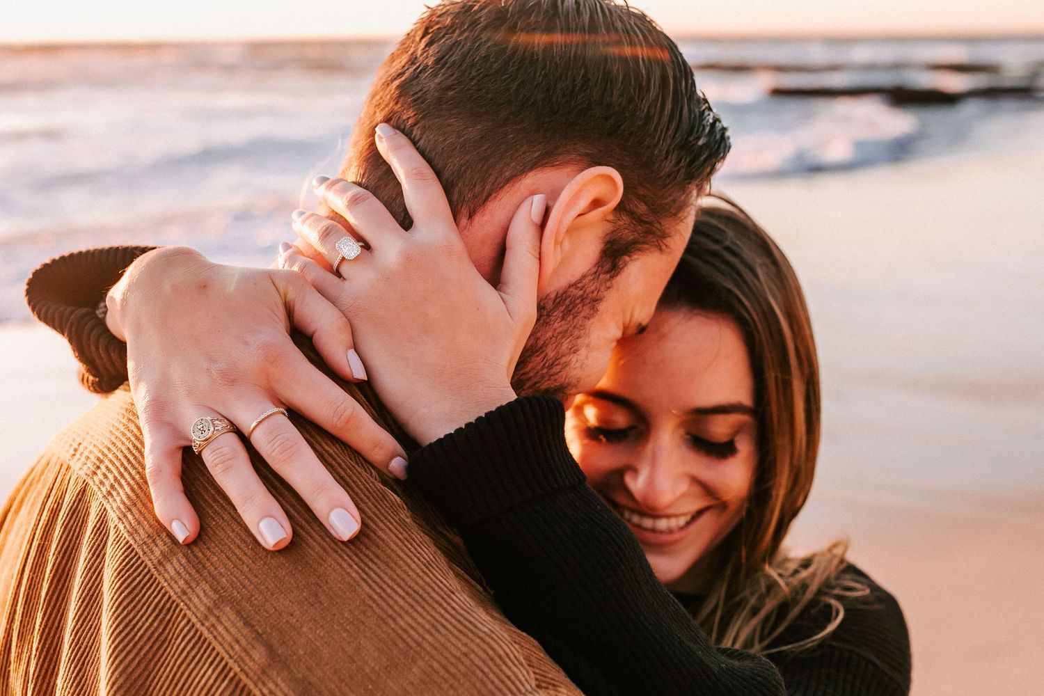 Close-up of smiling woman hugging man on beach, showing off engagement ring at sunset.