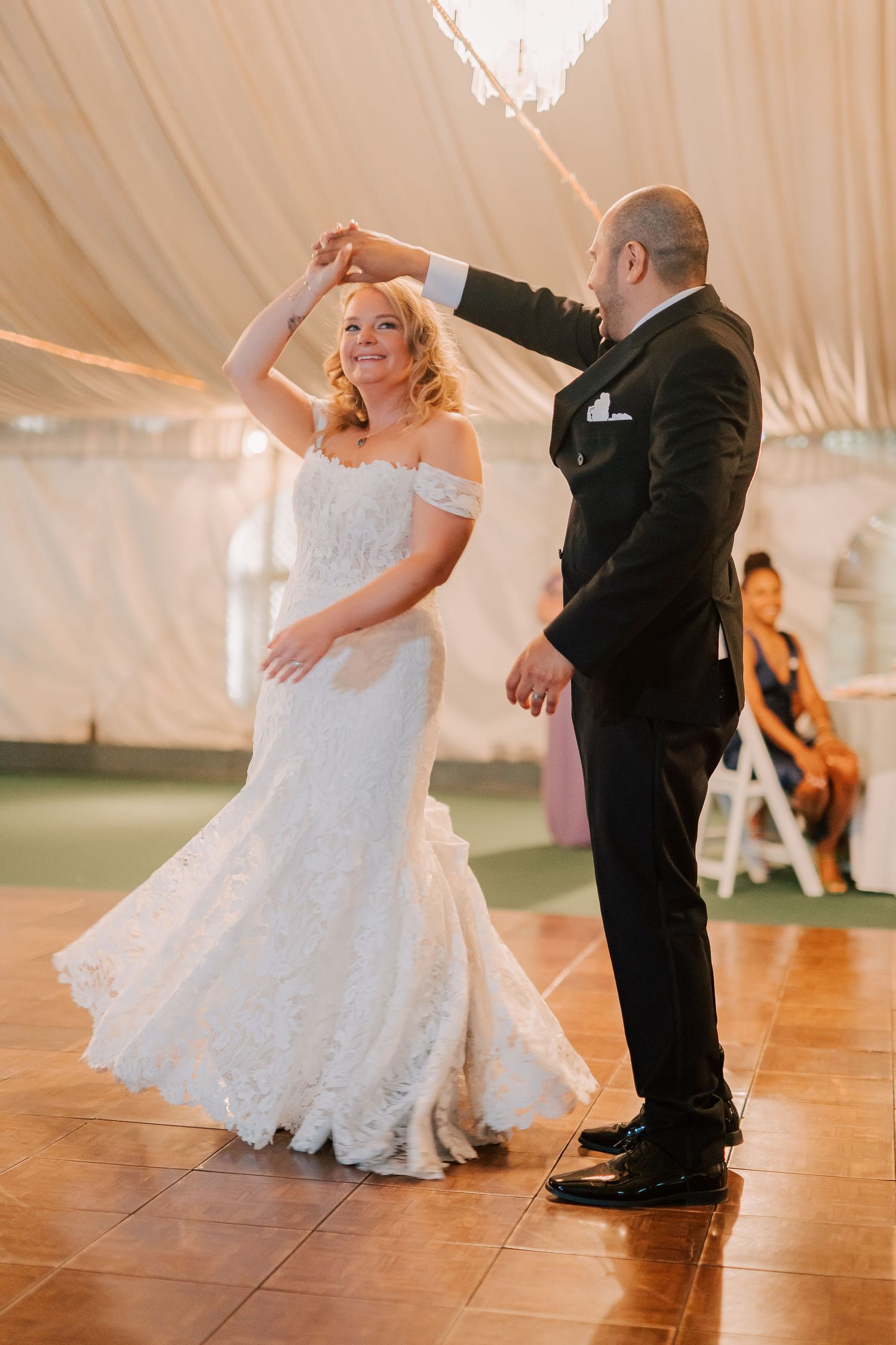 Bride and groom share their first dance under a tent, as groom twirls bride in a lace off-shoulder wedding gown.