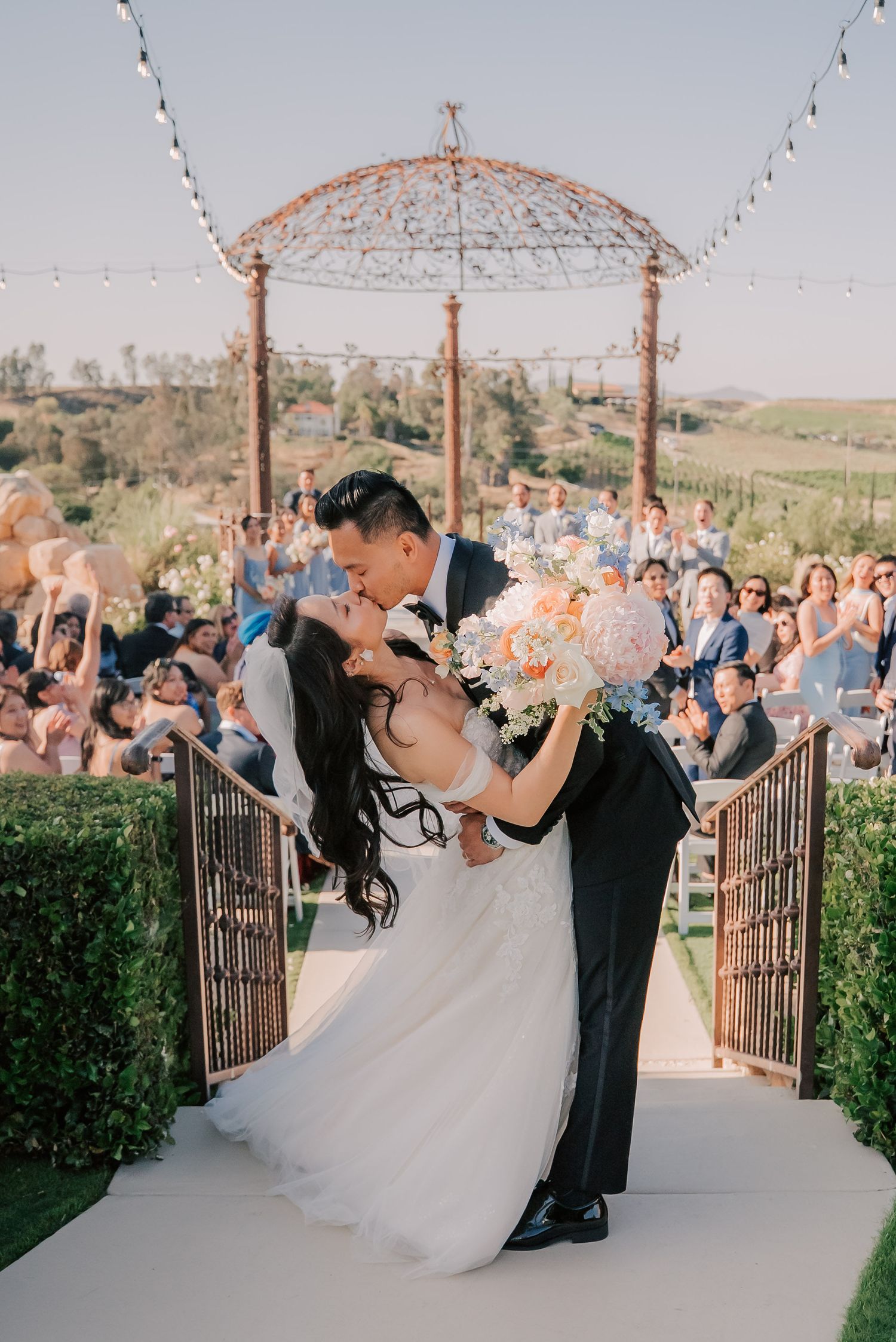 Bride and groom share a romantic kiss at outdoor vineyard wedding ceremony with guests cheering in background.