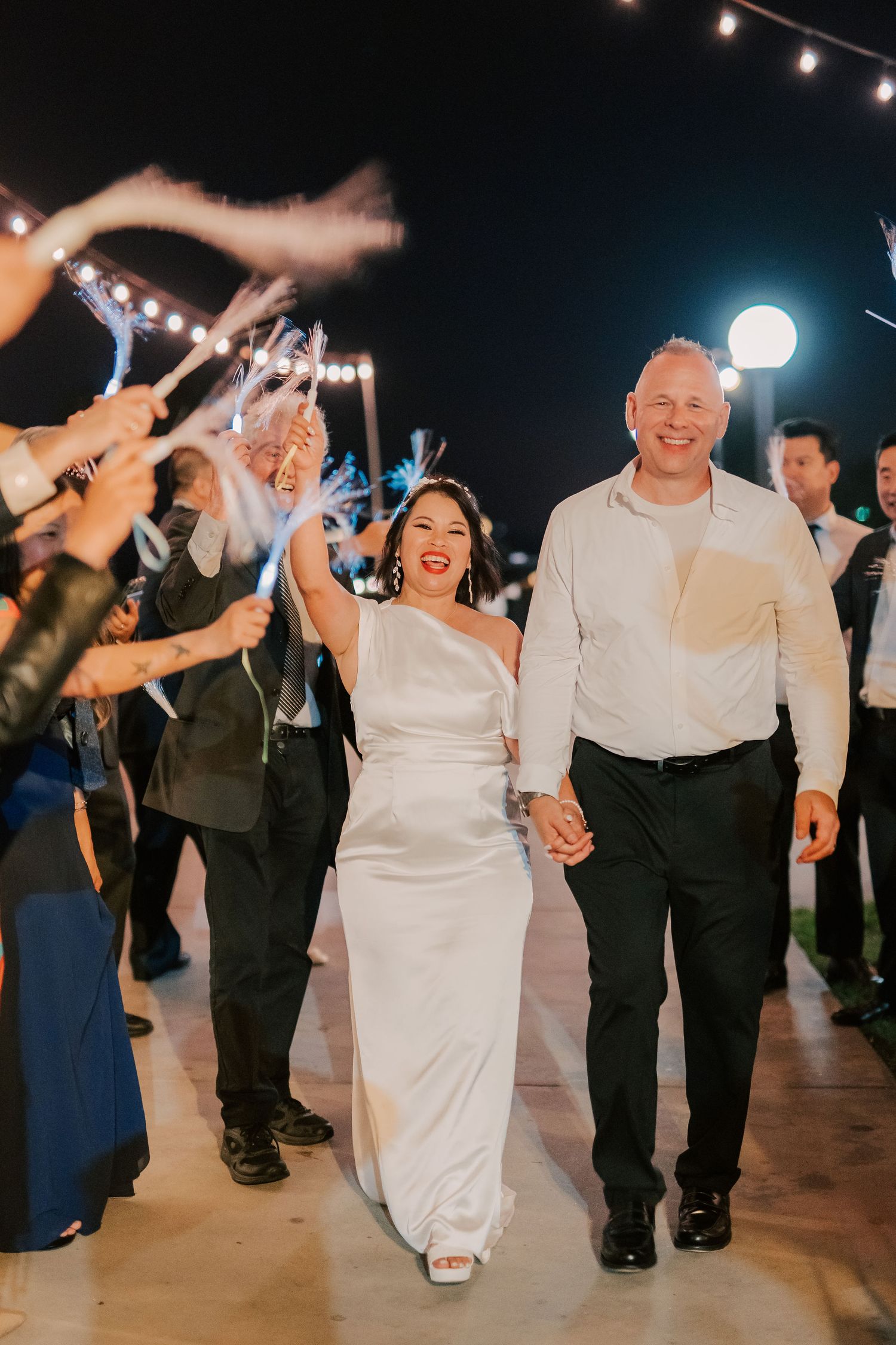 Bride and groom walk through a sparkler exit at their outdoor night wedding reception, smiling joyfully.
