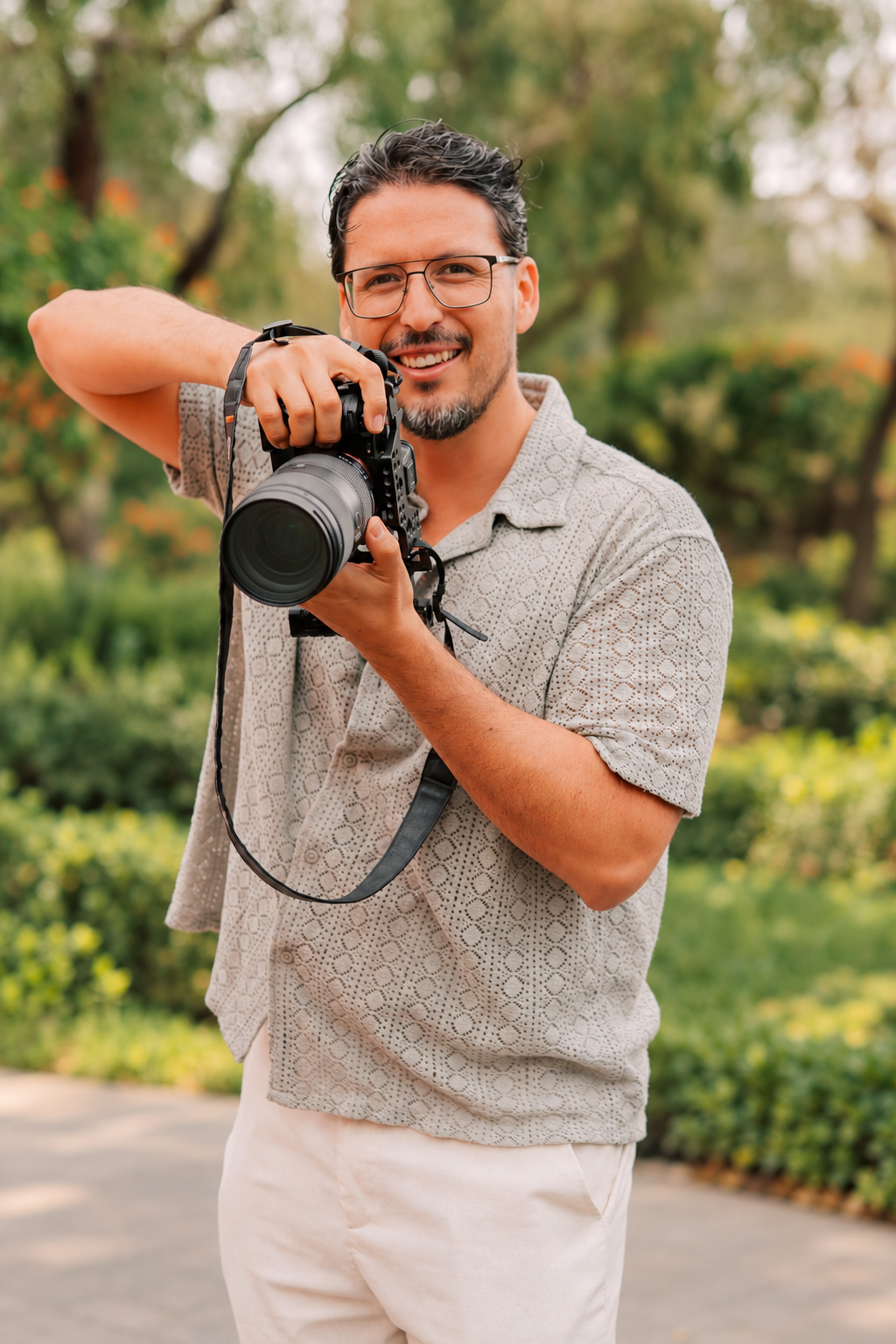 Smiling man with glasses holding a DSLR camera outdoors in a lush green park setting.