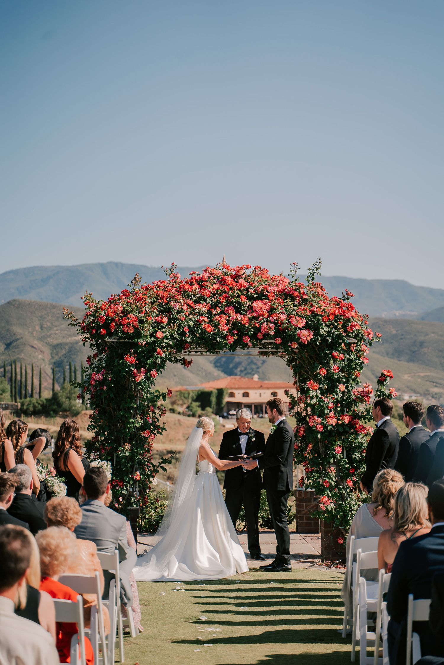 Outdoor wedding ceremony under a lush floral arch adorned with pink roses, set against a scenic mountain backdrop.