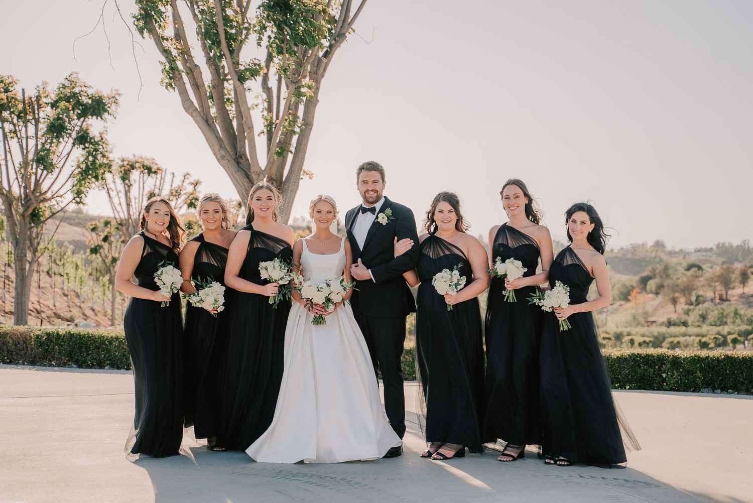 Bride and groom pose with six bridesmaids in black gowns holding white bouquets at outdoor wedding.