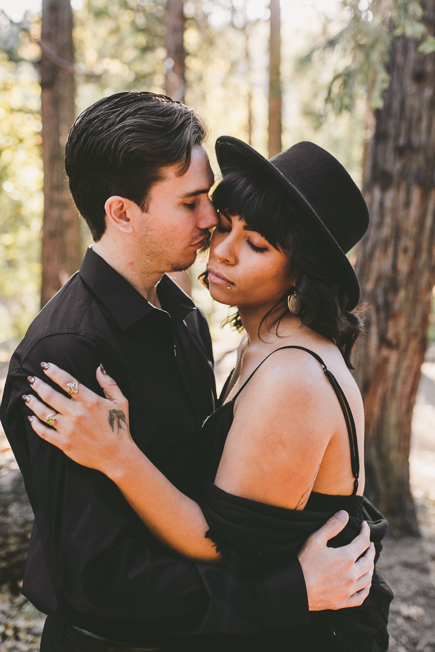 Couple in black outfits embracing in forest, woman wearing wide-brim hat, man kissing her forehead tenderly.