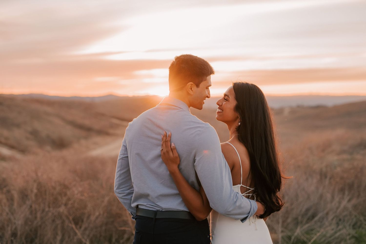 A couple embraces at sunset in a golden grassy field, the woman smiling over the man's shoulder as warm light glows on the horizon.