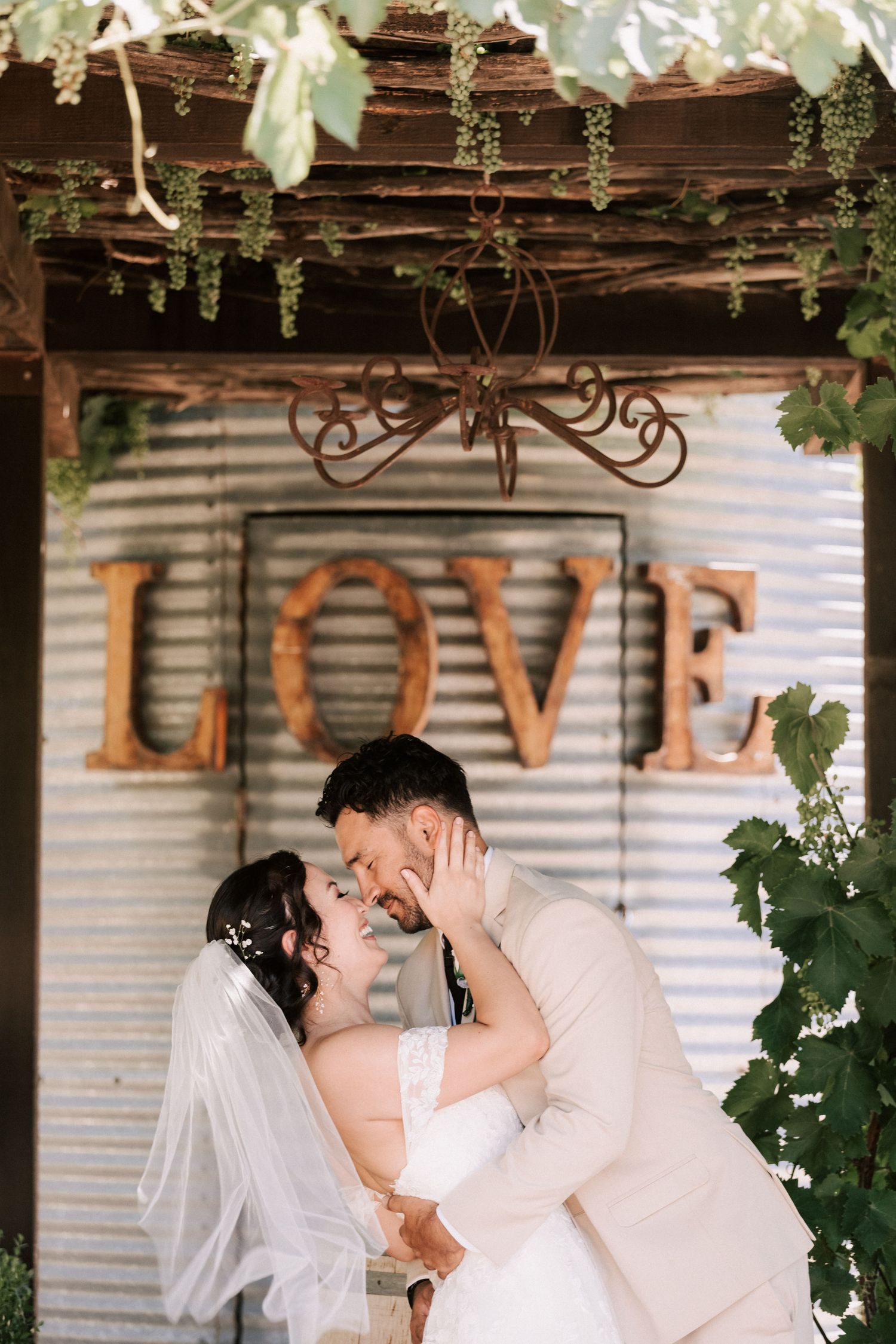 Newlyweds share a romantic kiss under a rustic LOVE sign surrounded by lush greenery at an outdoor wedding venue.