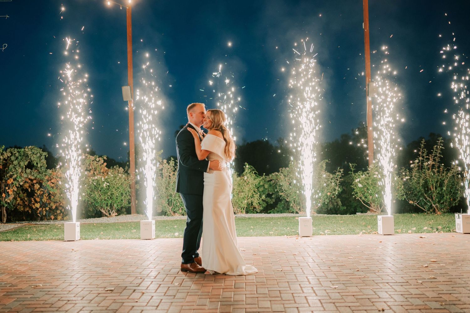 Bride and groom share romantic kiss surrounded by sparkling cold fireworks fountains at night wedding celebration.