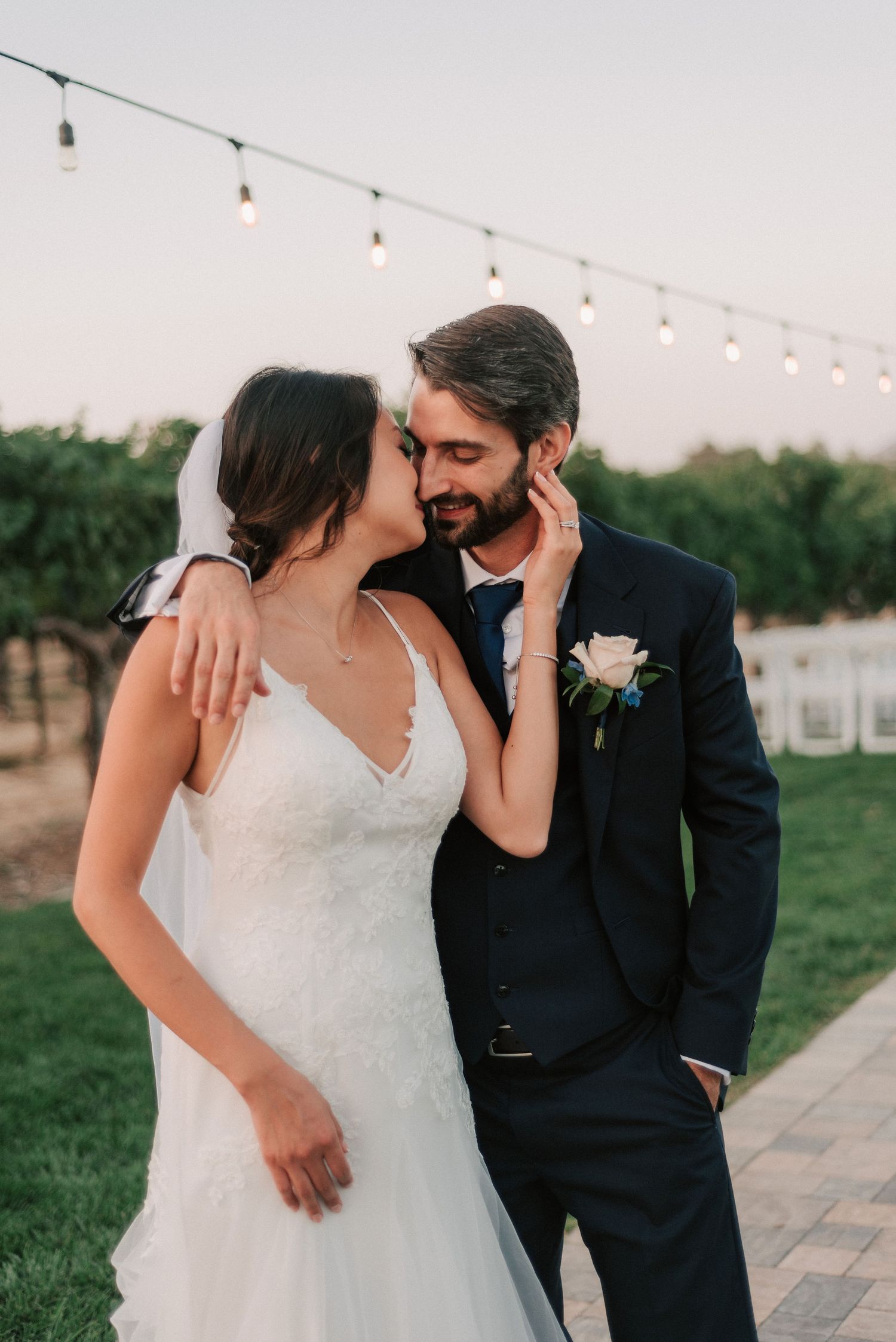 Bride in lace gown kisses groom in navy suit outdoors at vineyard wedding with string lights at dusk.
