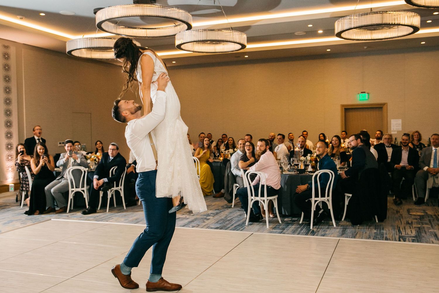 Groom lifts bride during first dance at elegant wedding reception with guests seated at round tables under modern chandeliers.