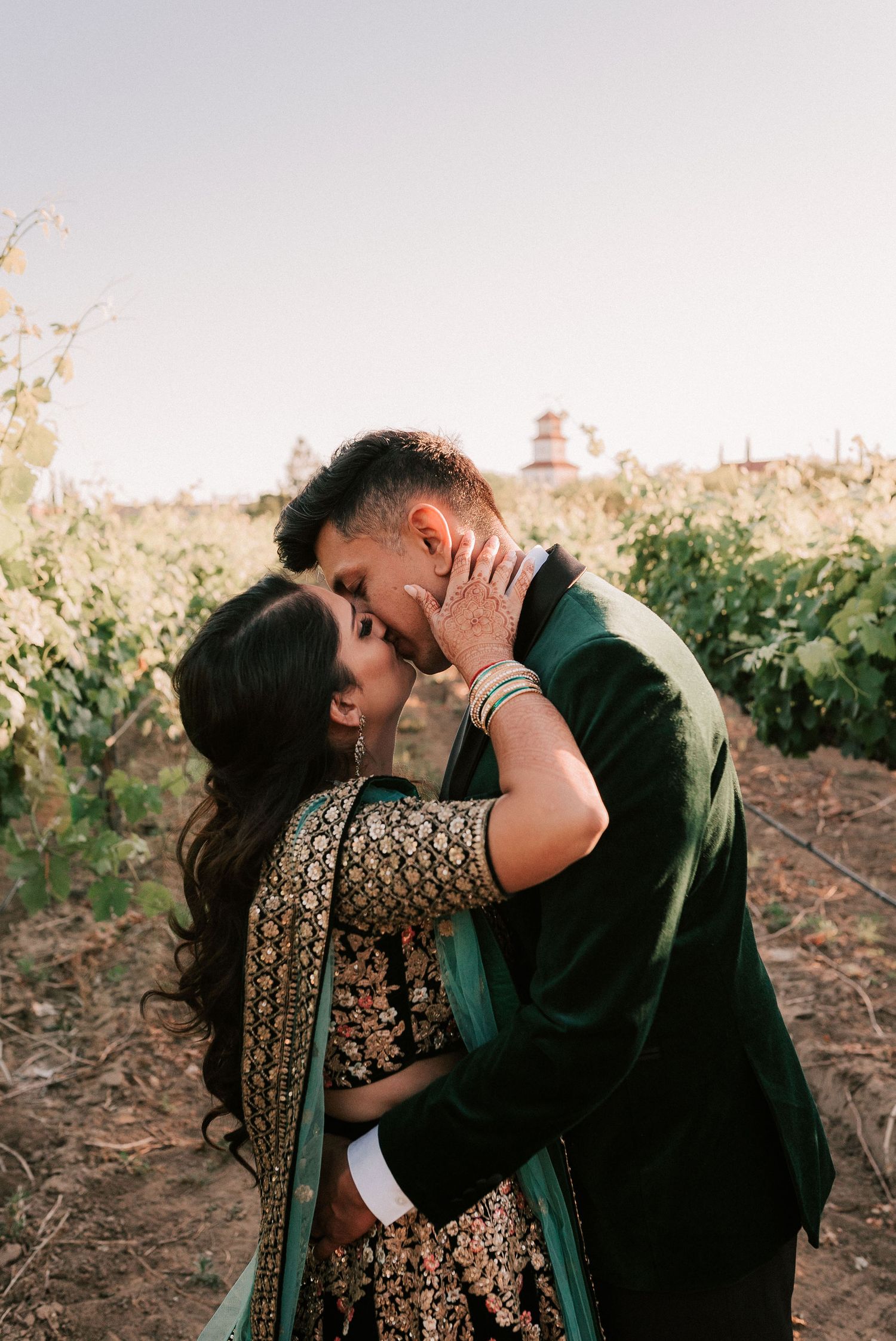 Couple shares romantic kiss in vineyard, bride wearing ornate green and gold Indian bridal attire.