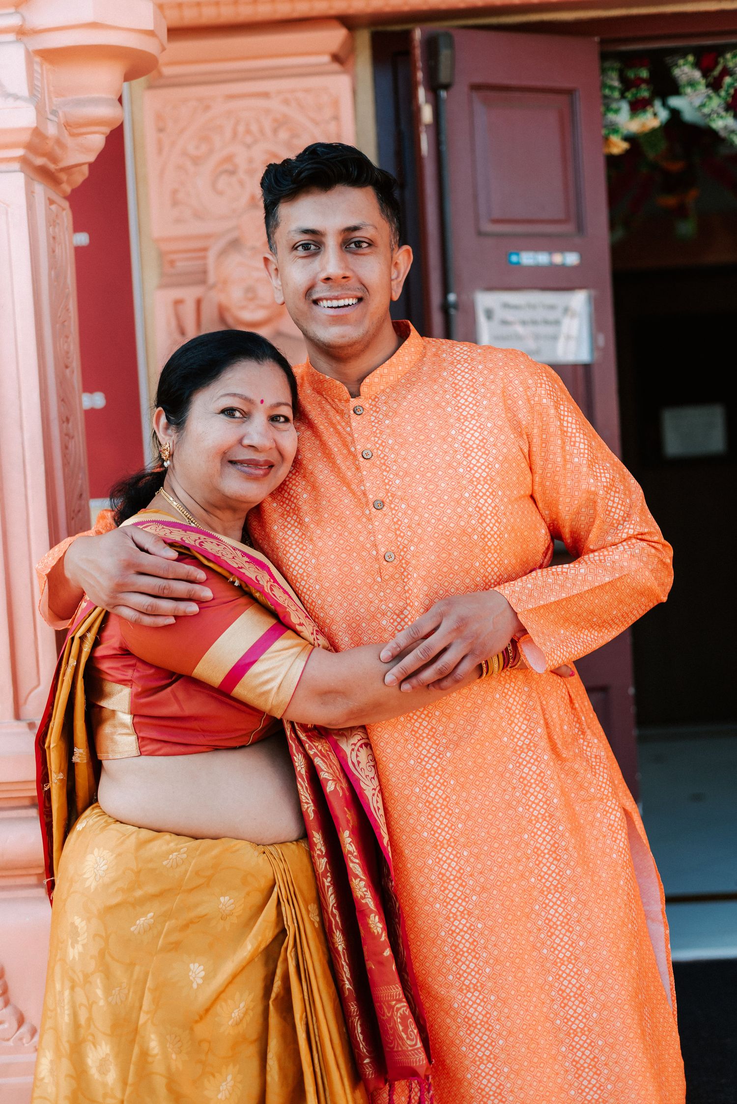 A smiling couple in traditional Indian attire, woman in yellow saree and man in orange kurta, posing at a temple.