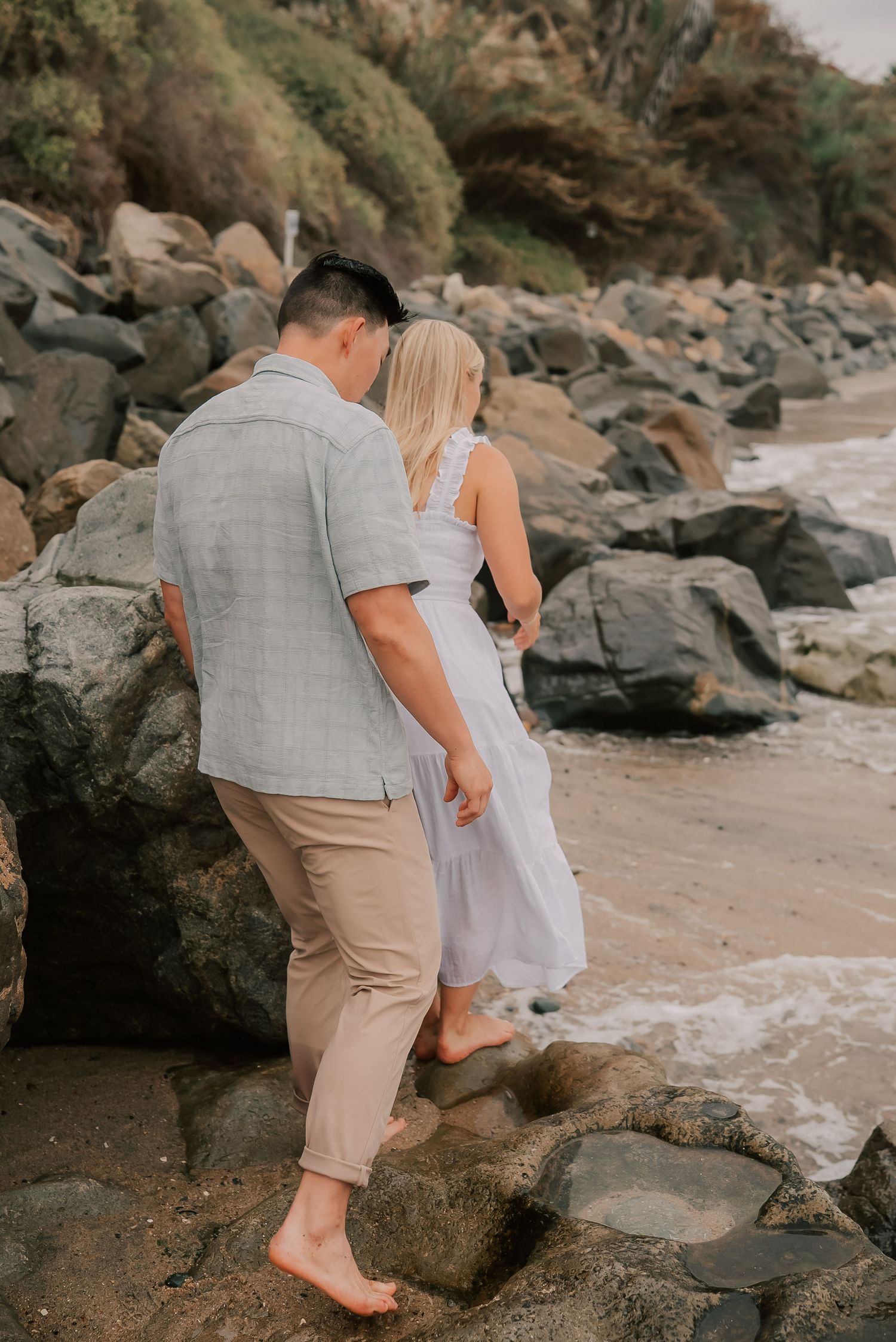 Couple walking barefoot on rocky beach, man in gray shirt and khakis, woman in white dress, ocean waves in background.