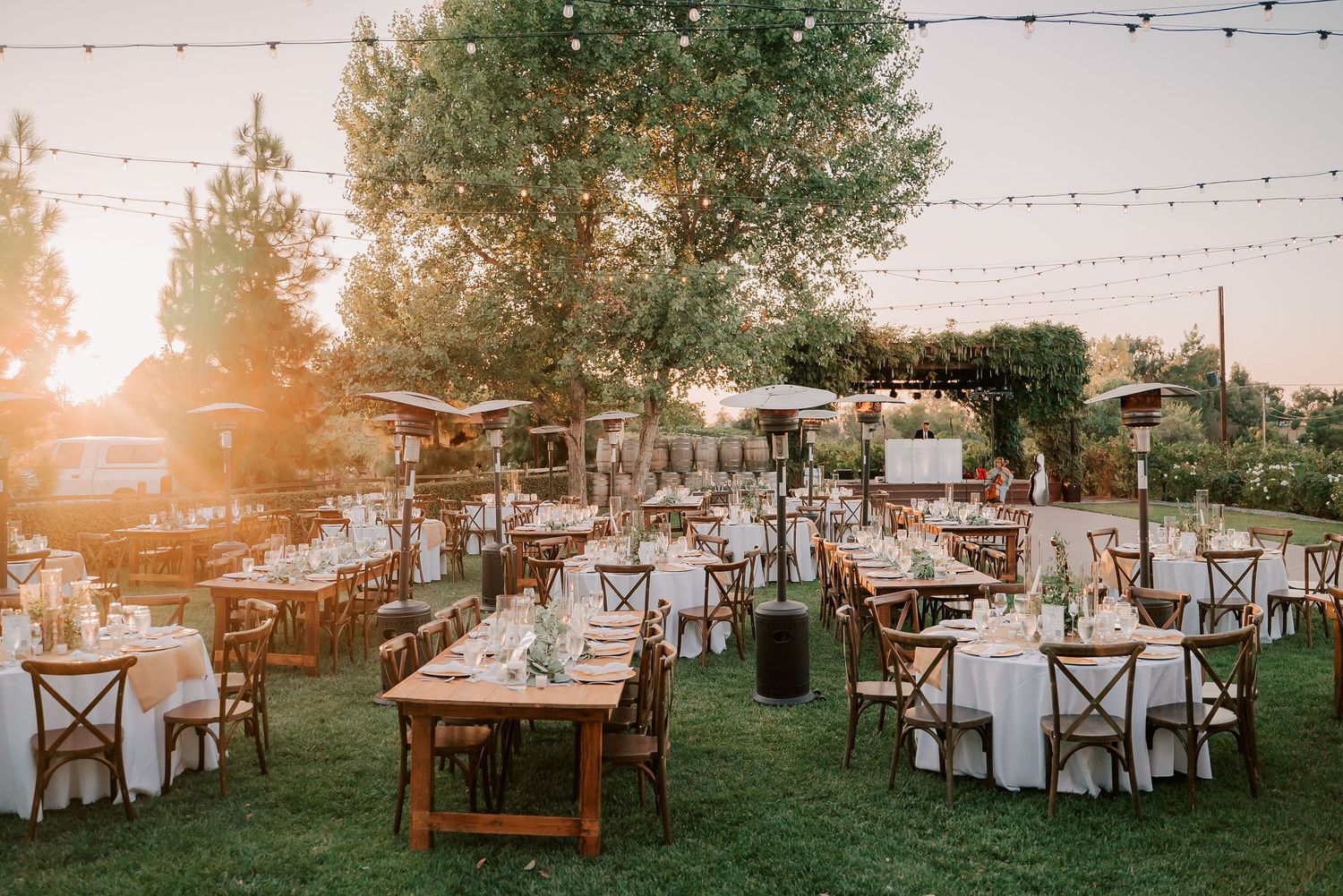 Outdoor wedding reception at golden hour with string lights, wooden tables, white linens, and lush greenery in a garden setting.