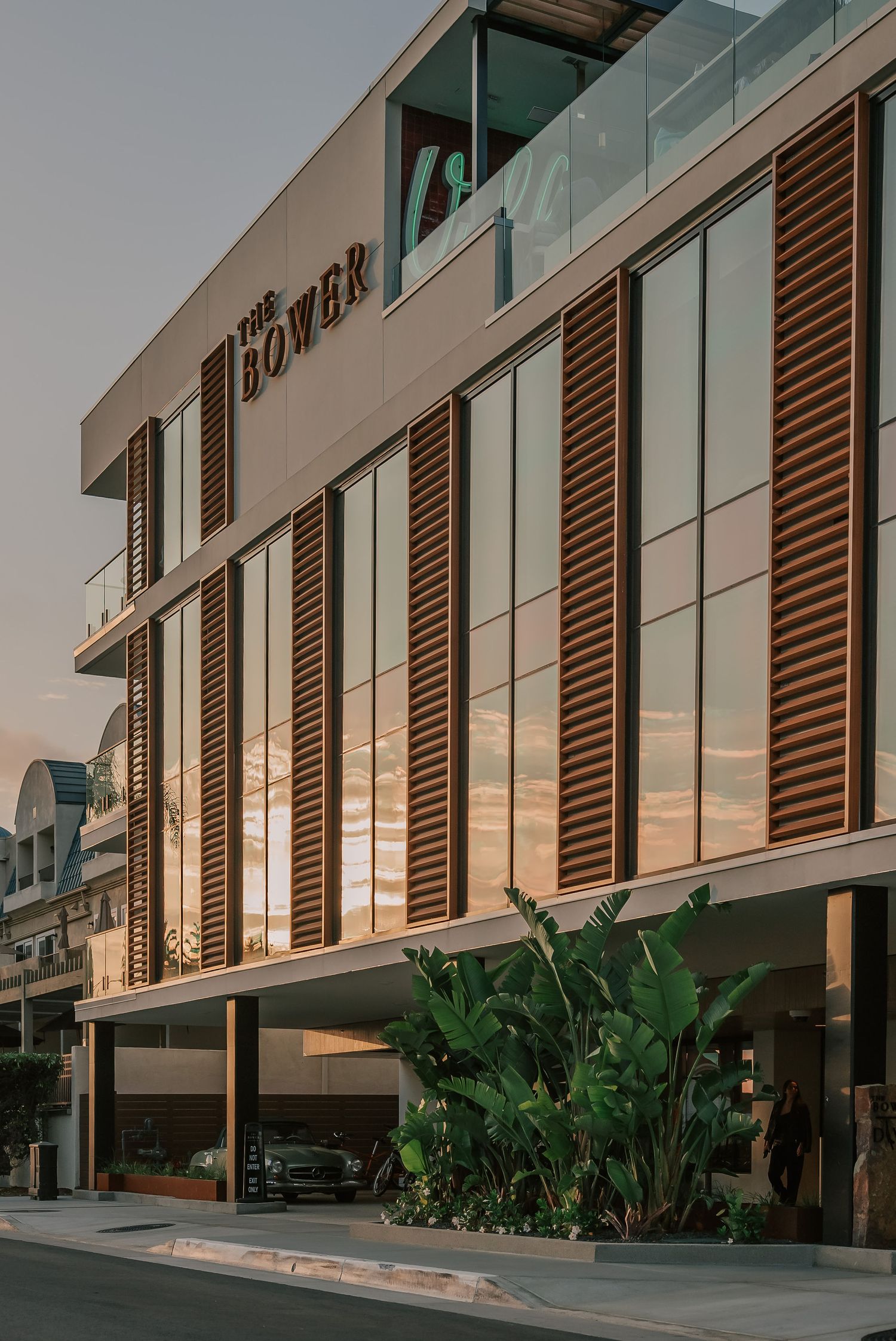The Bower building exterior featuring wooden louvers, glass facade, and tropical plants at golden hour.