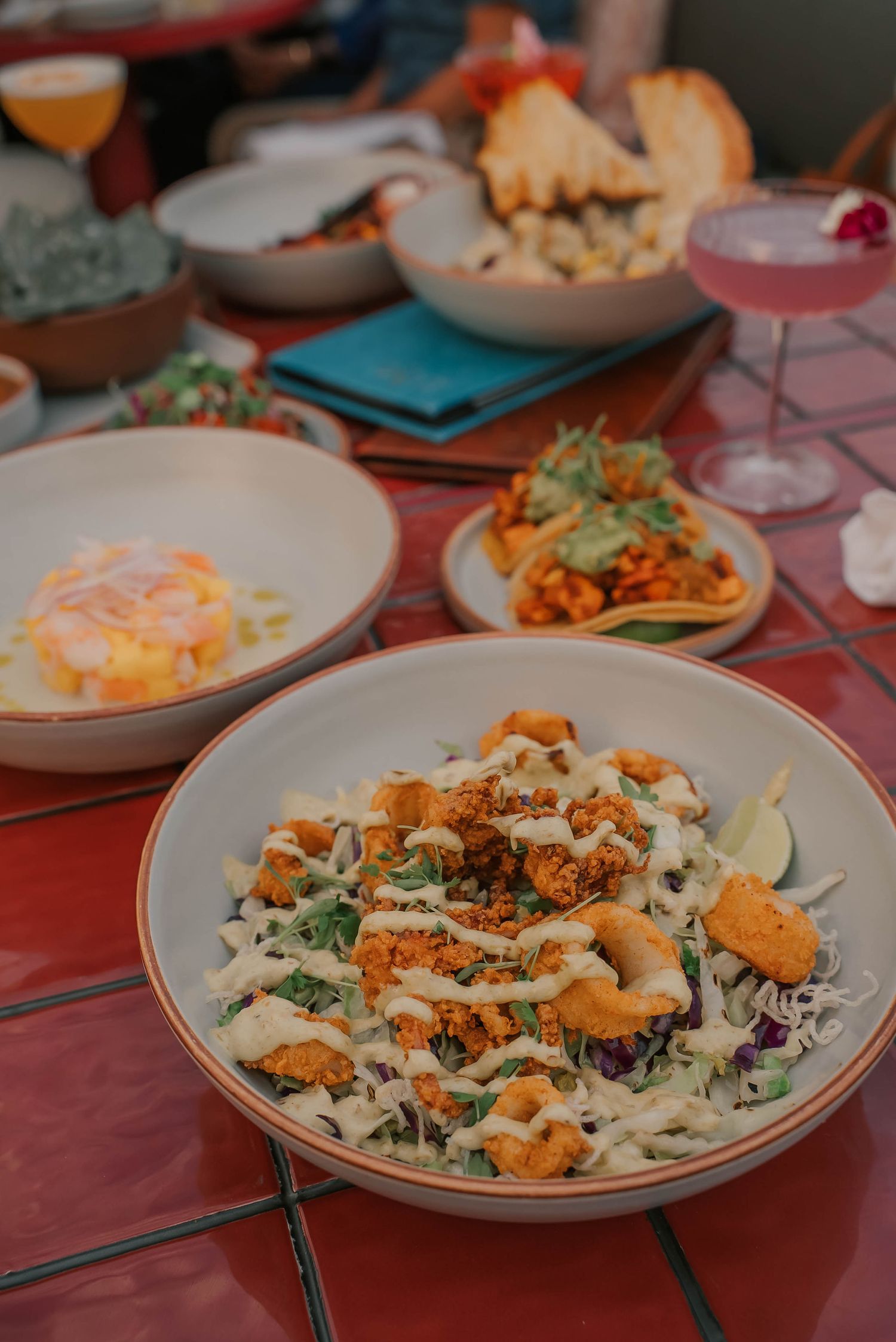 A vibrant spread of Mexican dishes on a red tile table, featuring a crispy chicken salad, tacos, and colorful cocktails.
