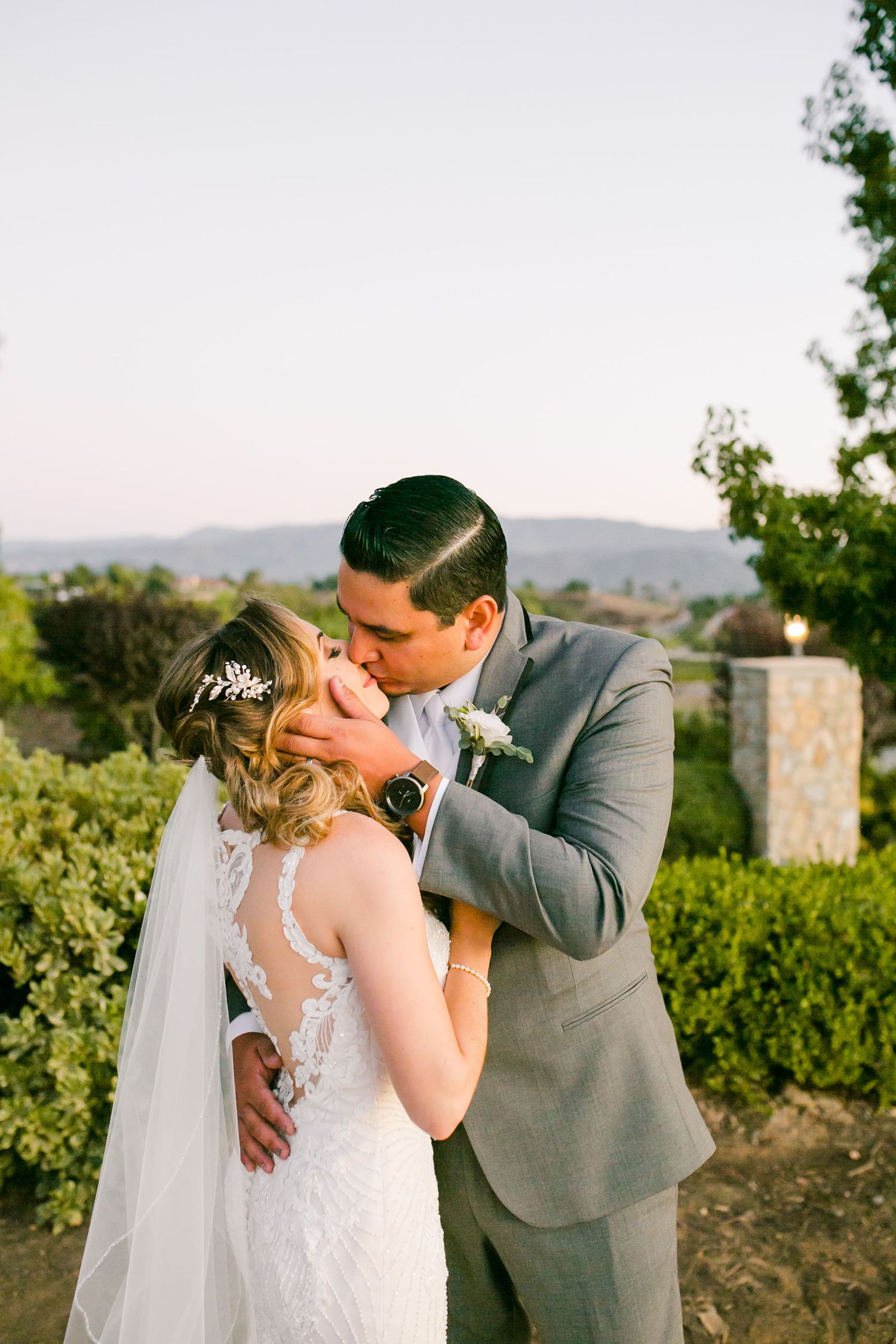 Bride and groom share a romantic kiss outdoors at golden hour, with rolling hills and lush greenery in the background.