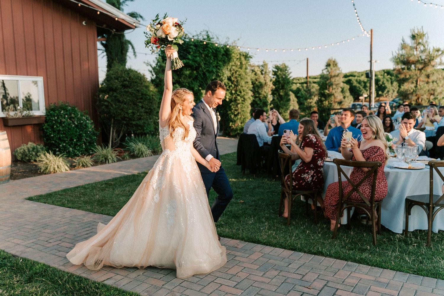 Bride raises bouquet while groom escorts her past guests at an outdoor wedding reception on a brick pathway.