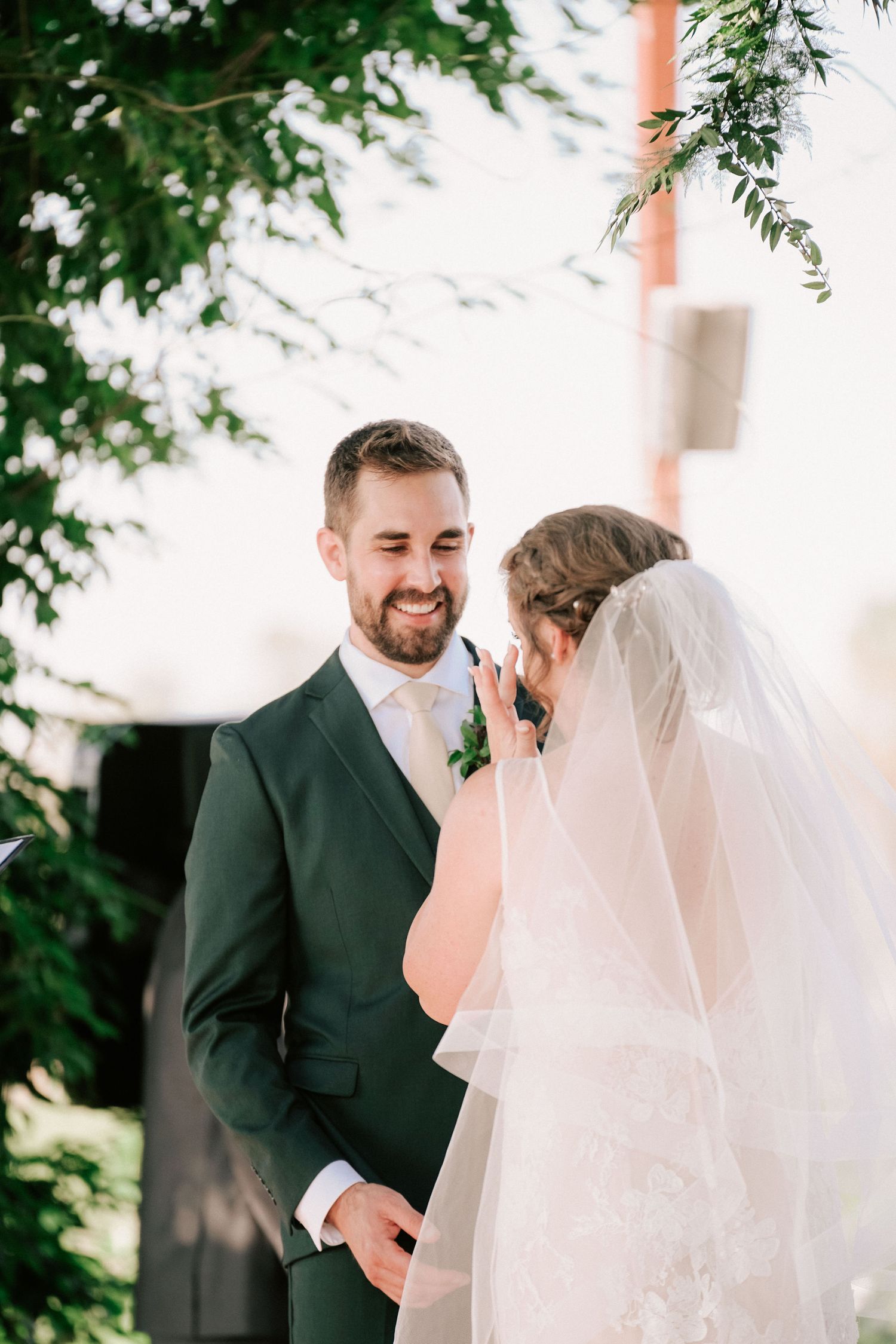 Groom in dark green suit smiling at bride in white wedding dress and veil during outdoor ceremony.