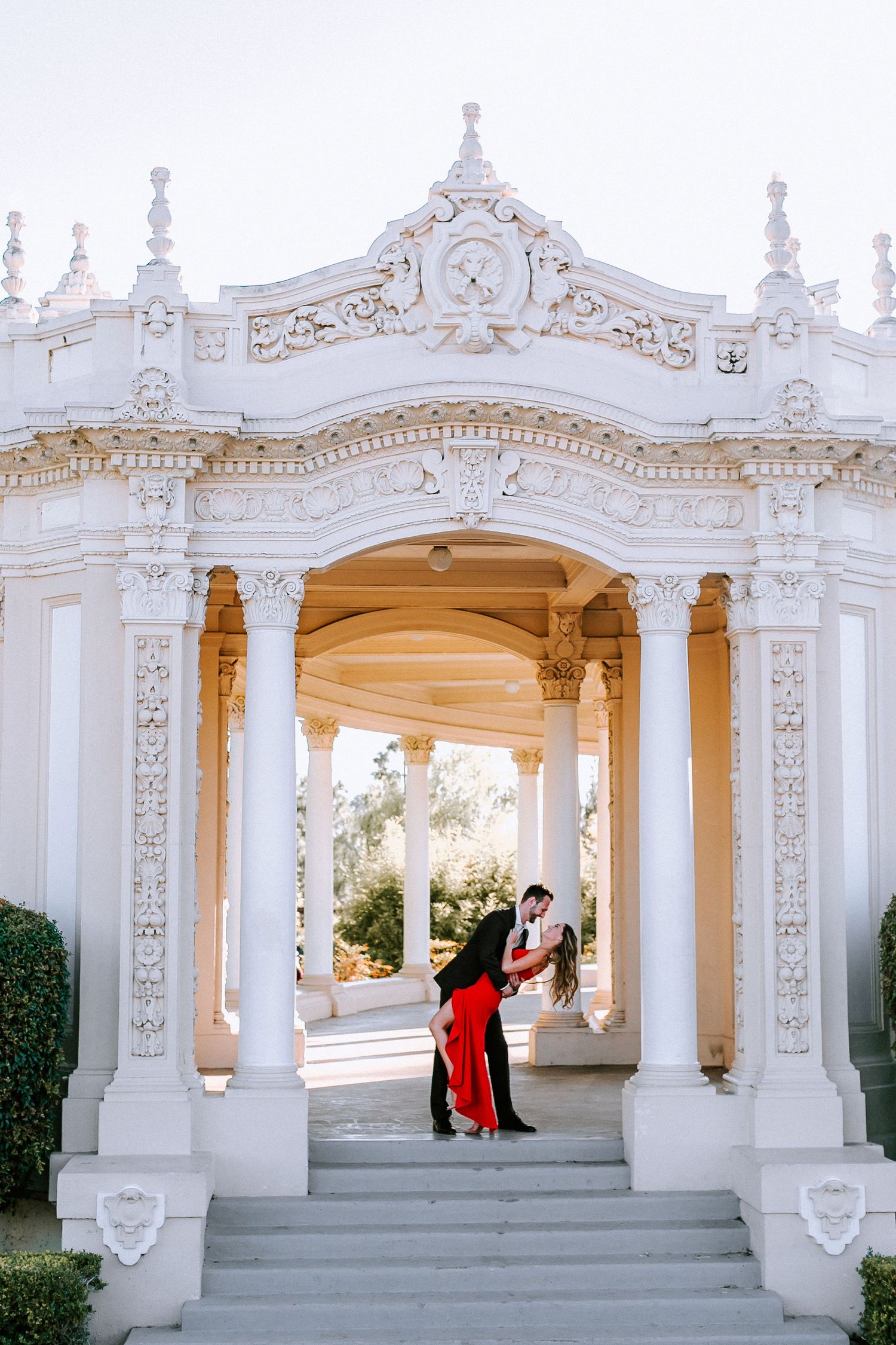 Couple embracing romantically under ornate white arched colonnade with elegant architectural details.
